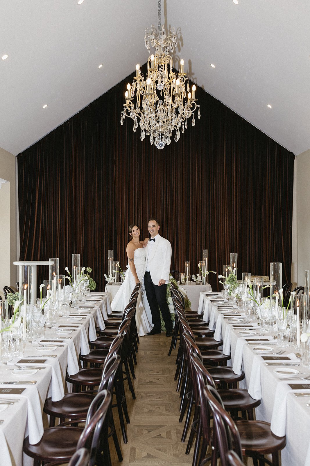 A newlywed couple in wedding attire stands in the center of a decorated banquet hall, with a large chandelier hanging from the ceiling. The hall features long tables on either side, set with white tablecloths, glassware, and floral centerpieces. Dark