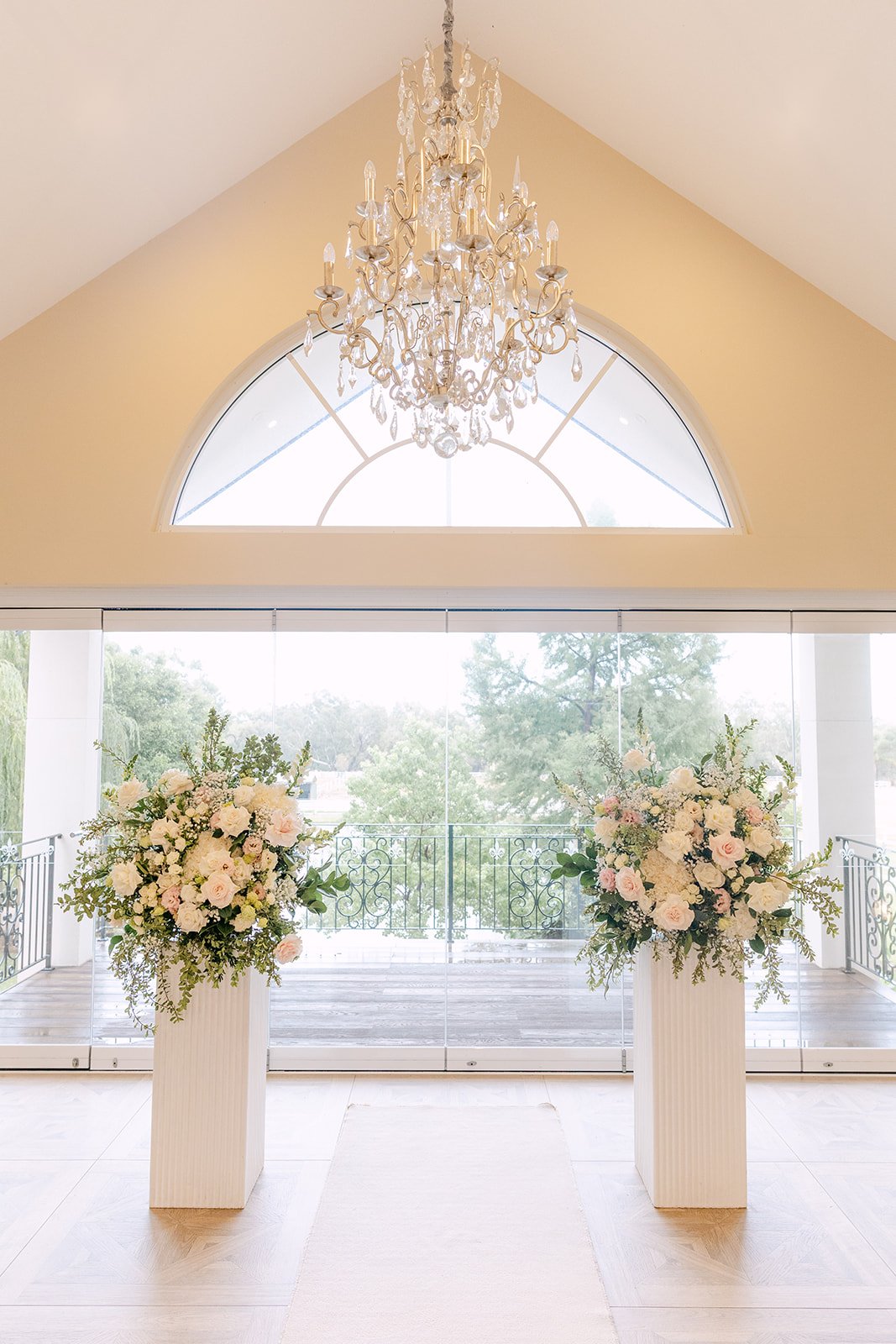 Elegant home interior with large arched window, crystal chandelier, and floral arrangements on pedestals.