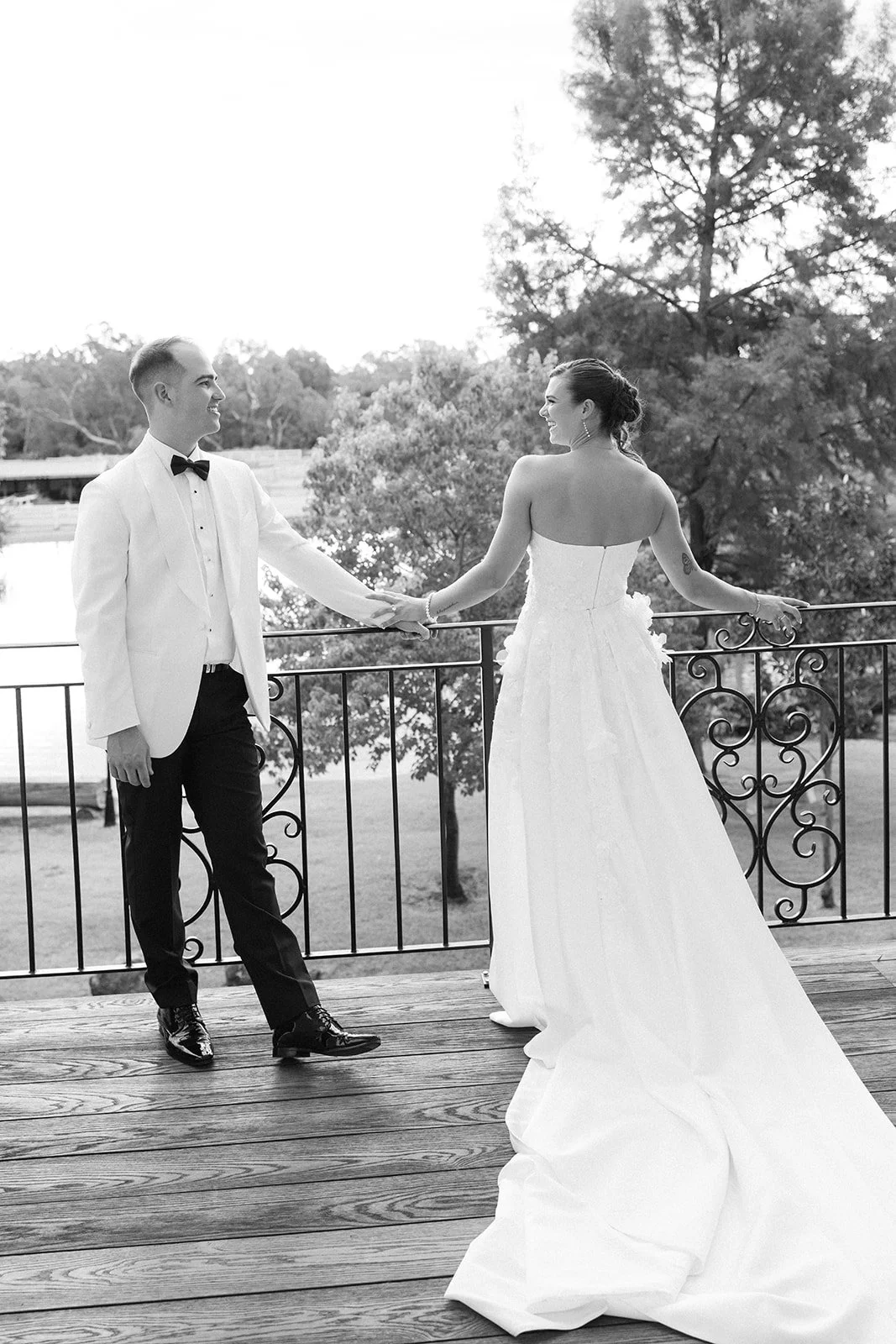 A black and white photo of a bride and groom holding hands on a balcony. The groom wears a tuxedo with a bow tie, and the bride is in a strapless wedding gown with a long train. They are smiling at each other with trees and water in the background.