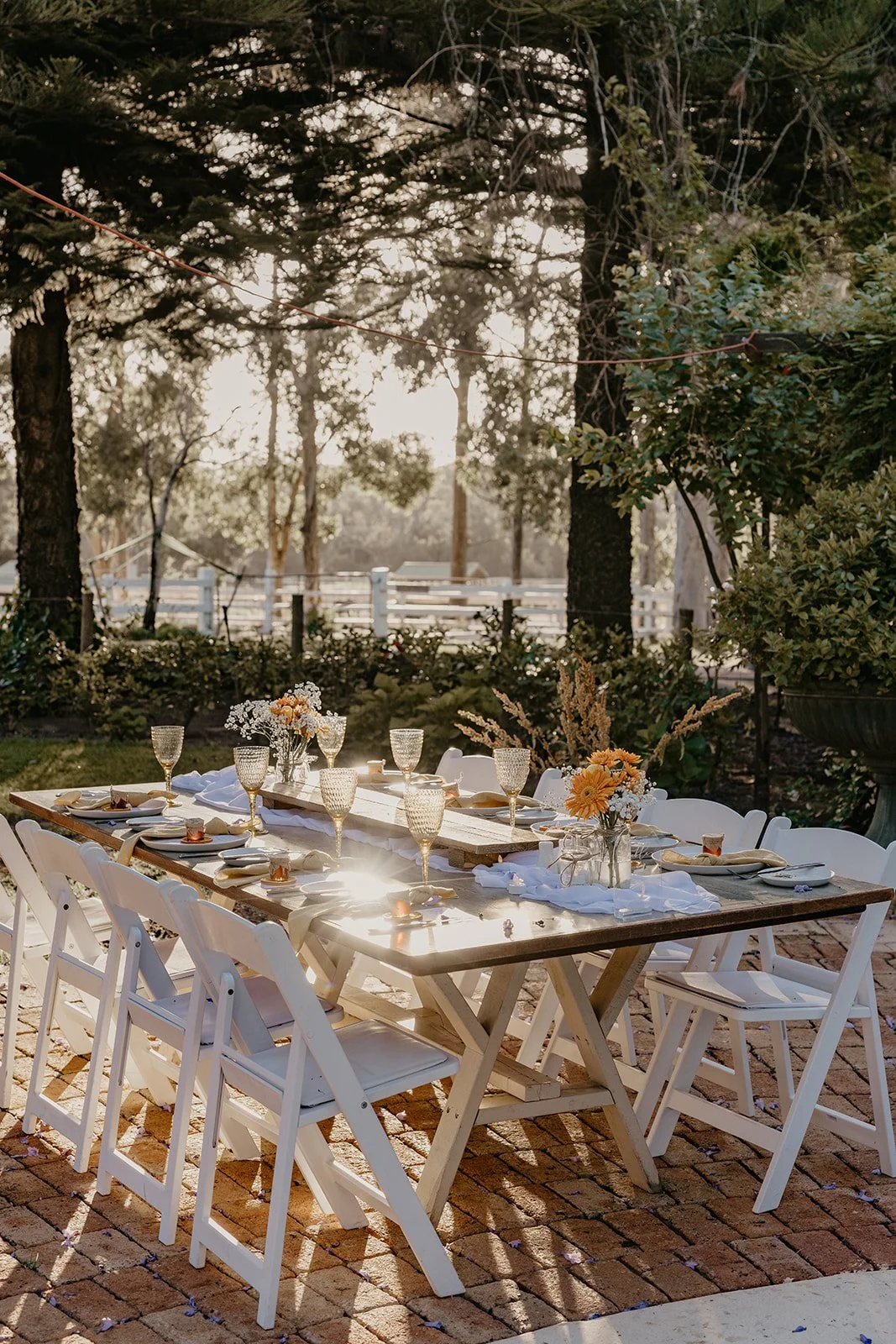 Outdoor dining table set with white chairs, glassware, plates, and floral centerpieces, on a brick patio with trees and sunlight in the background.