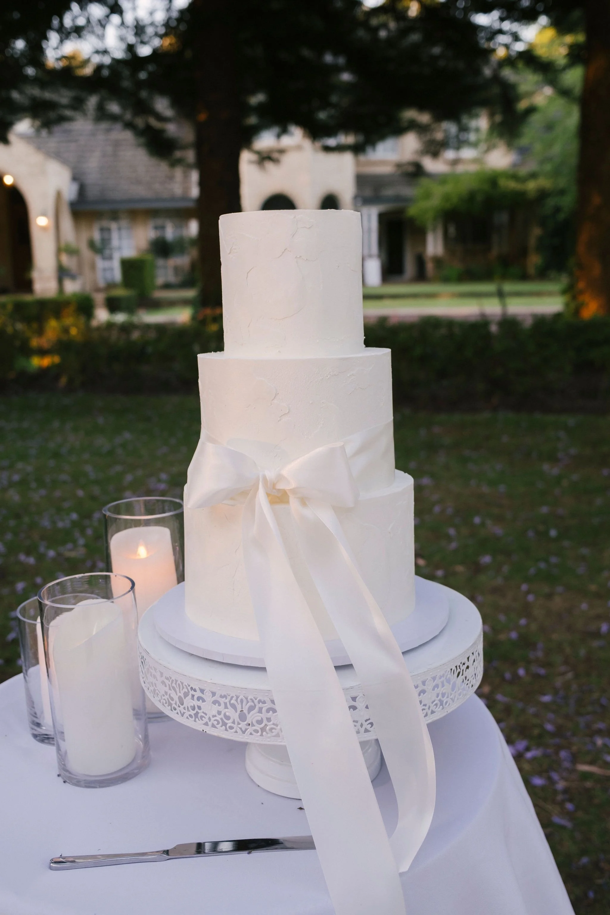 A three-tier white wedding cake with textured icing, decorated with a white satin ribbon tied in a bow, placed on a white cake stand outdoors. There are two white candles in glass holders and a cake knife on the table. The background shows trees and houses, indicating an outdoor setting.