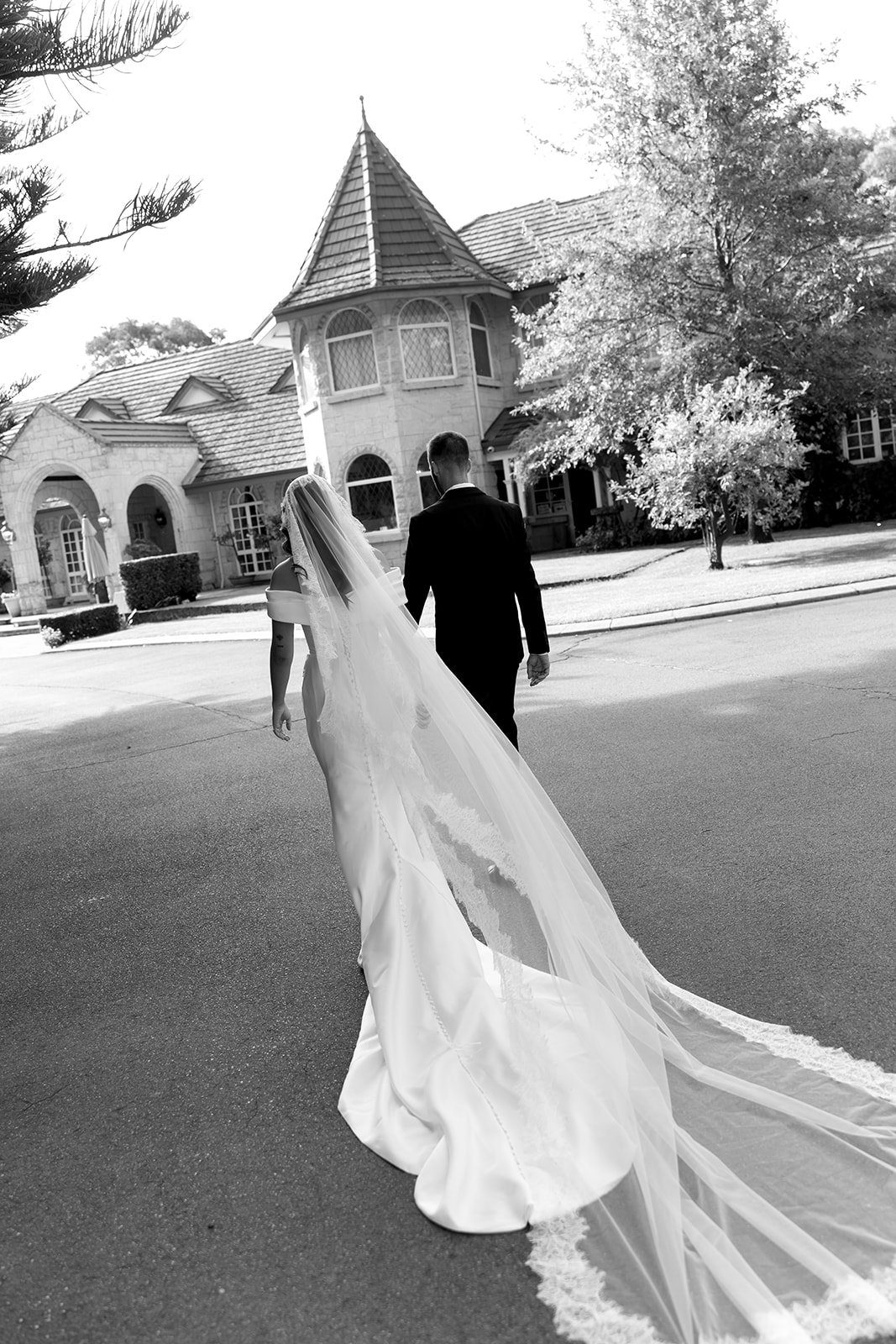 A bride and groom walking hand in hand down a quiet street in front of a large, ornate house with a tower-like structure on a sunny day.