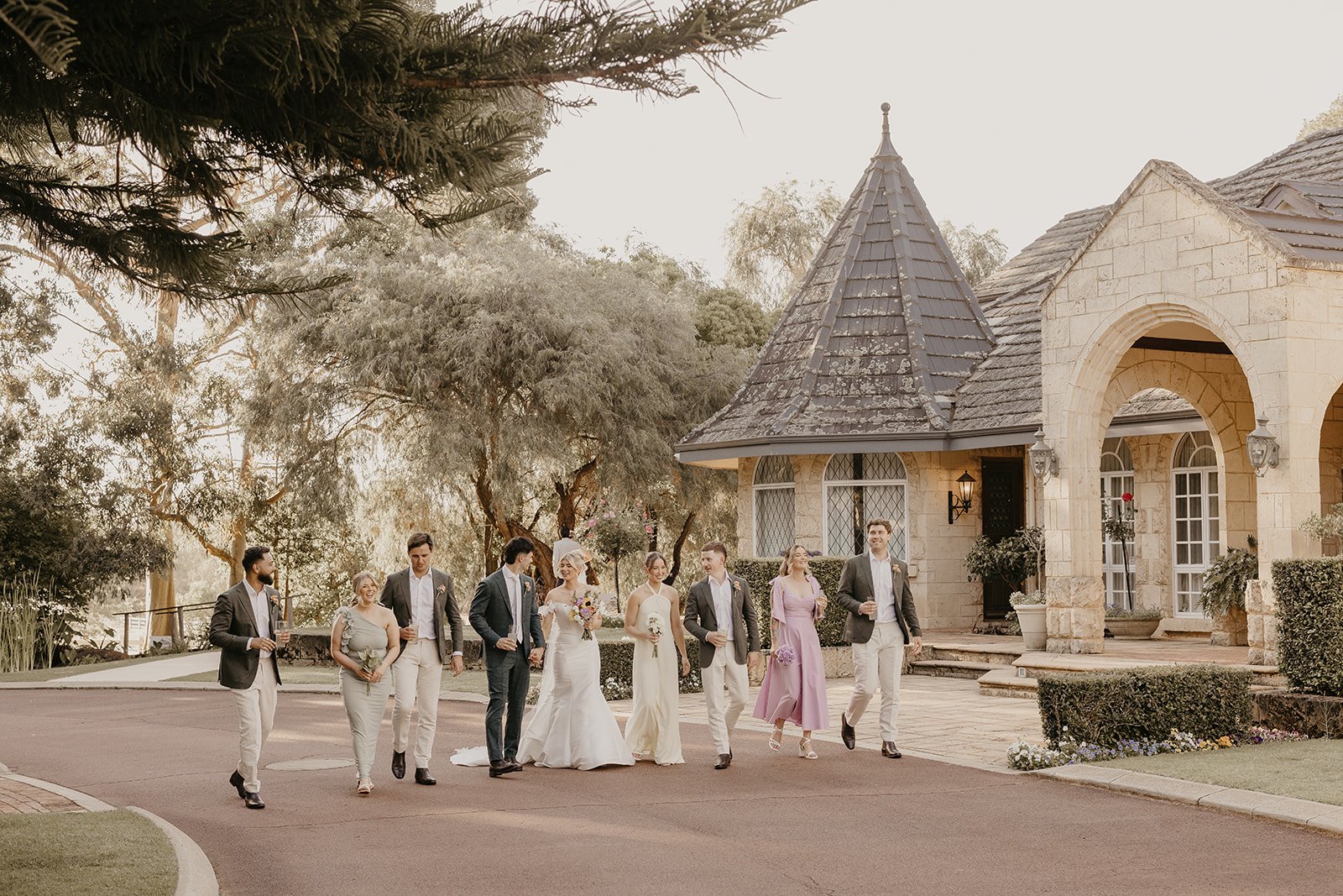 A bride in a white gown and a groom in a suit walk with wedding party outside a stone building with a conical roof, surrounded by trees and greenery.