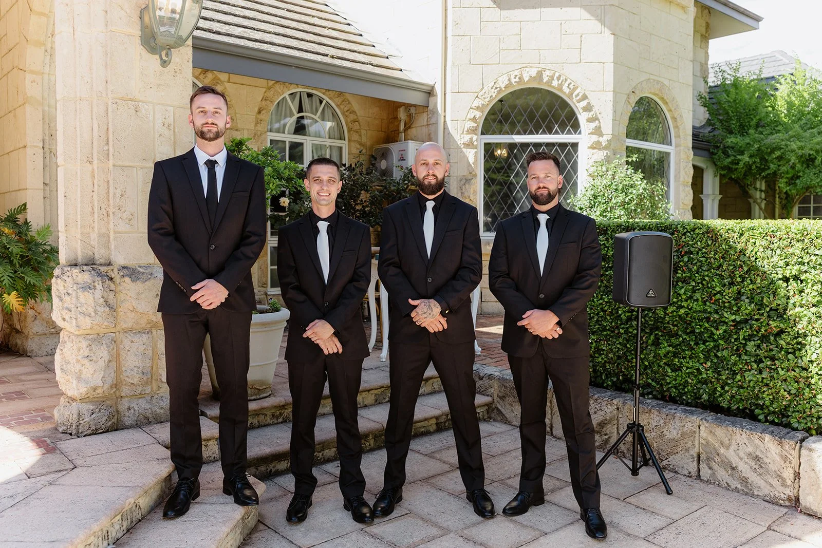 Four men in black suits and white ties standing on stone steps outside a house with beige stone walls and large arched windows. There is a speaker on a stand to the right and greenery surrounding the area.