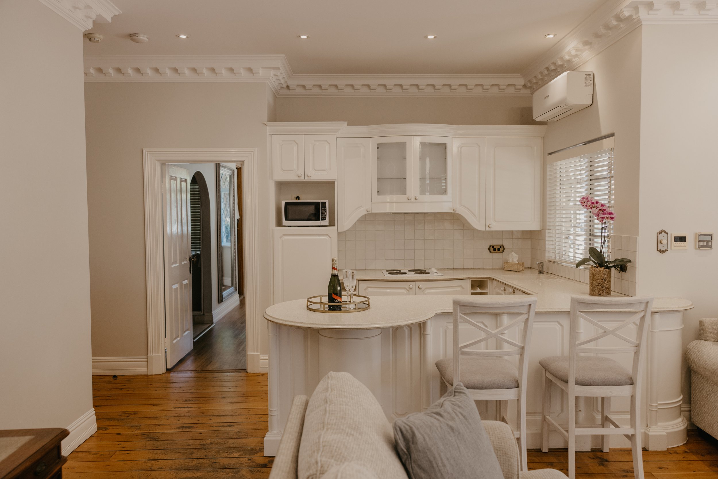 A kitchen with white cabinets, a beige countertop, a small microwave, a window with blinds, and a potted orchid on the counter. There is a round table with chairs and bag of snacks on a tray, and a doorway leading to another room.