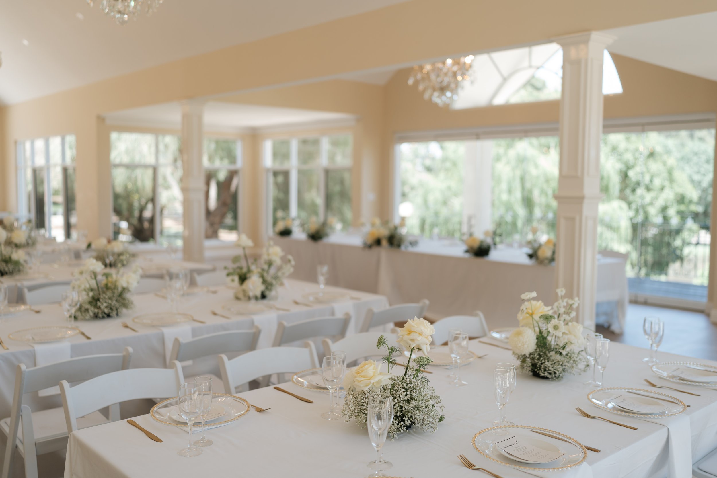 A decorated banquet hall set for a formal event, with long white tables covered in white tablecloths, adorned with floral centerpieces of white and cream flowers, and surrounded by white chairs. Large windows let in natural light, and chandeliers han