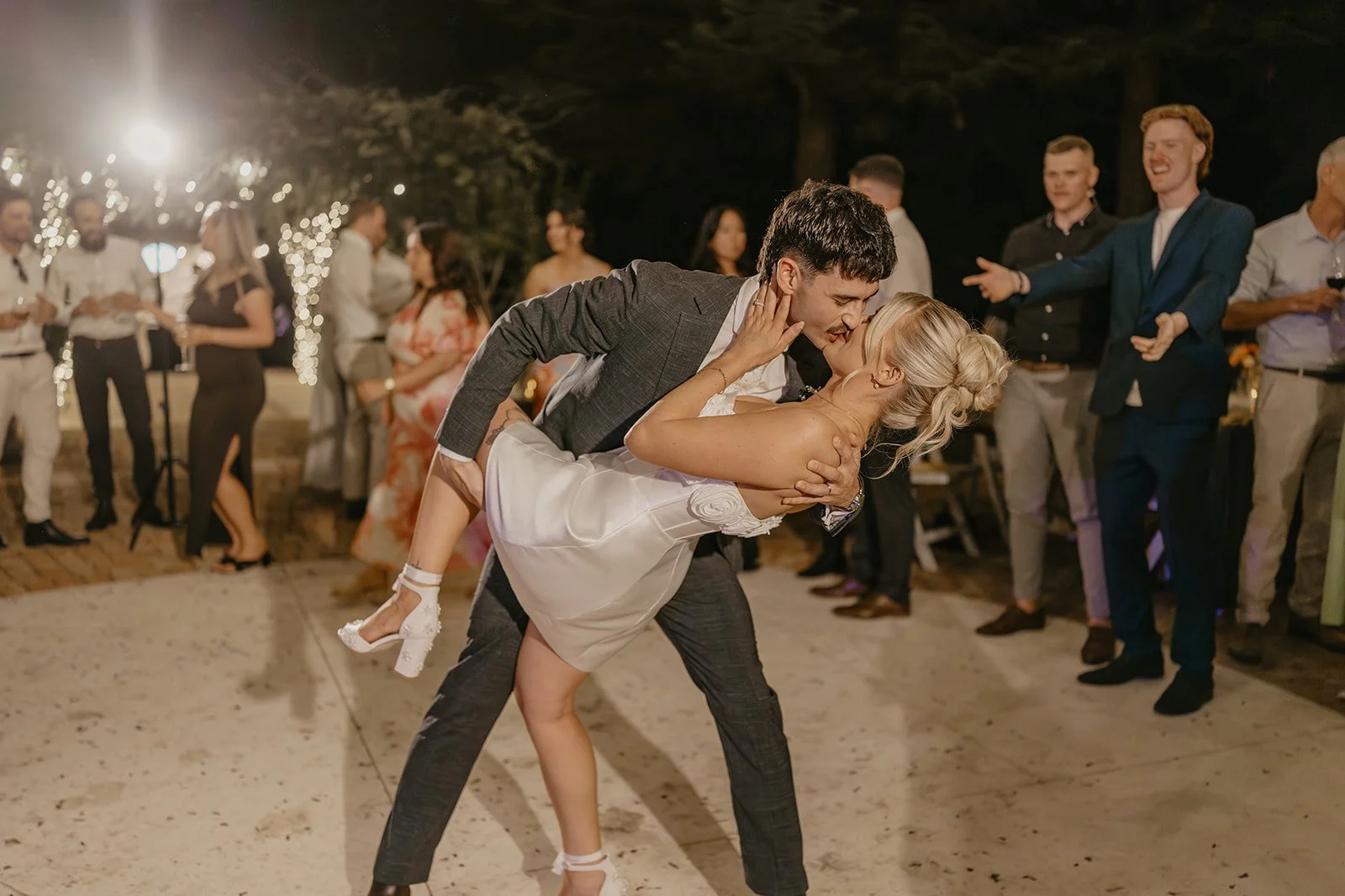 A man in a suit dancing with a woman in a white dress, dipping her backward at a celebration or wedding. People are watching and smiling in the background under outdoor string lights at night.