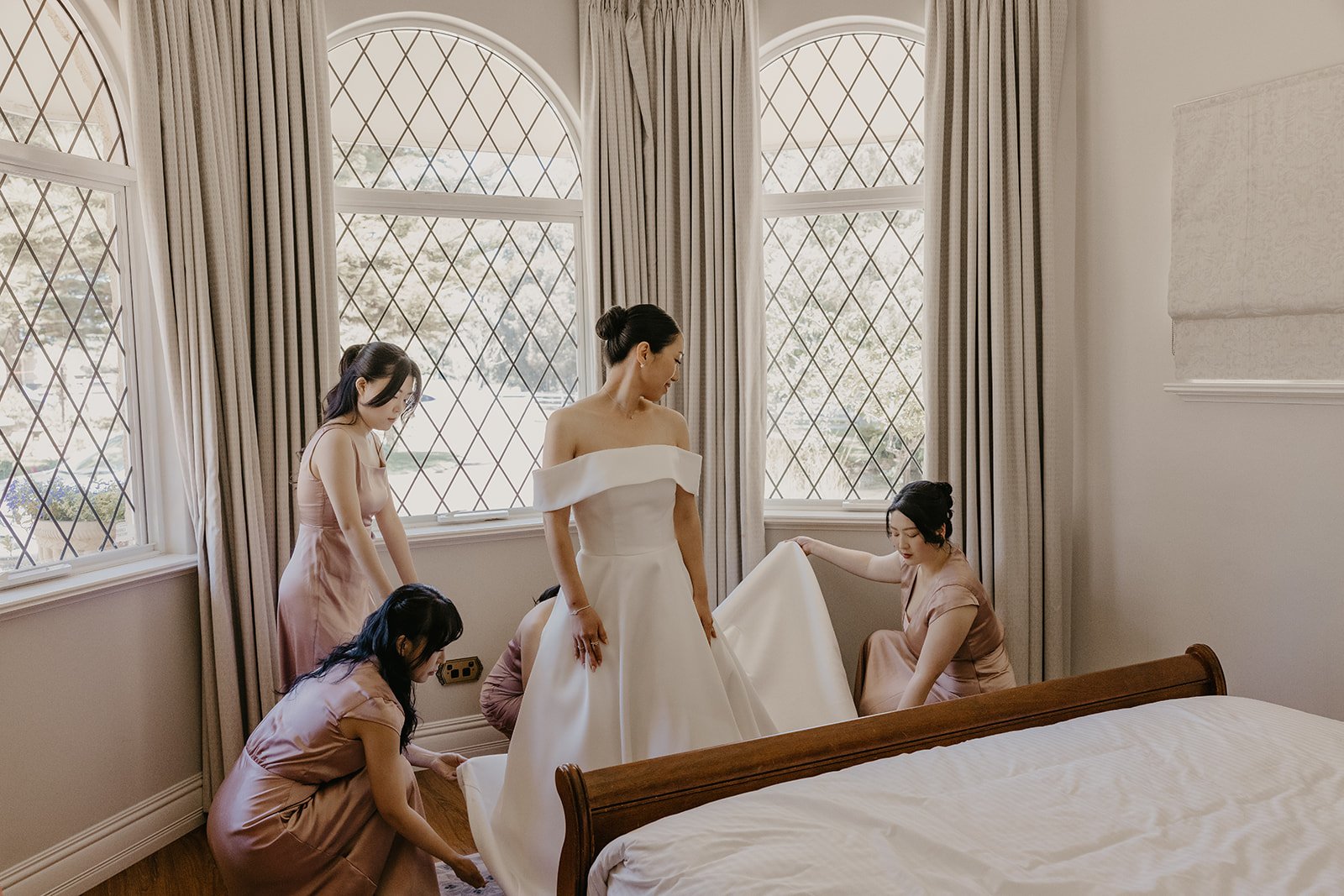 Women in a room with large, arched windows. One woman in a white dress is standing while four women in pink dresses are helping her with her gown.