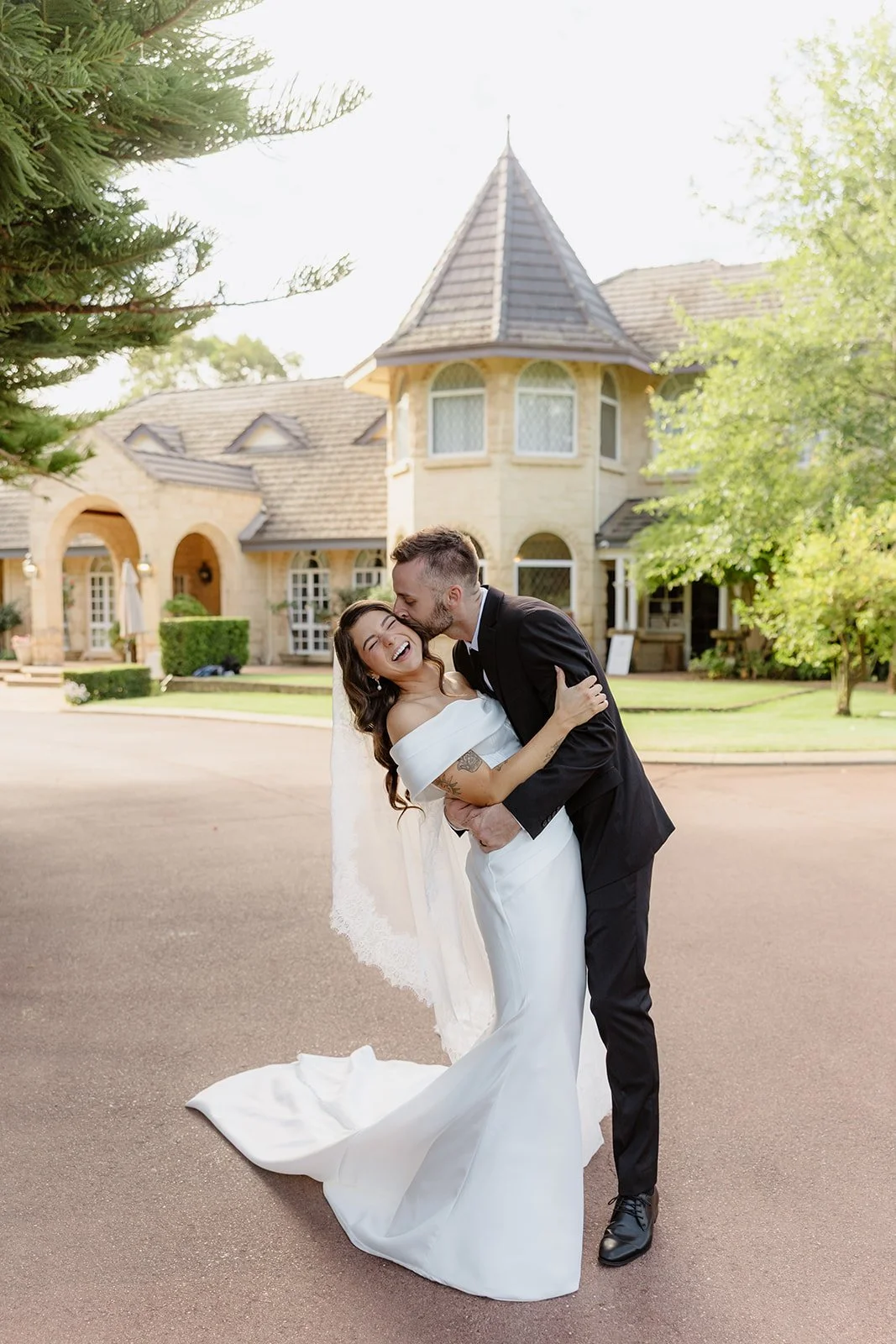 A newlywed couple sharing a joyful moment outside a large, elegant house, with the groom kissing the bride on the forehead, and the bride laughing, in a lush green yard with trees and shrubs.