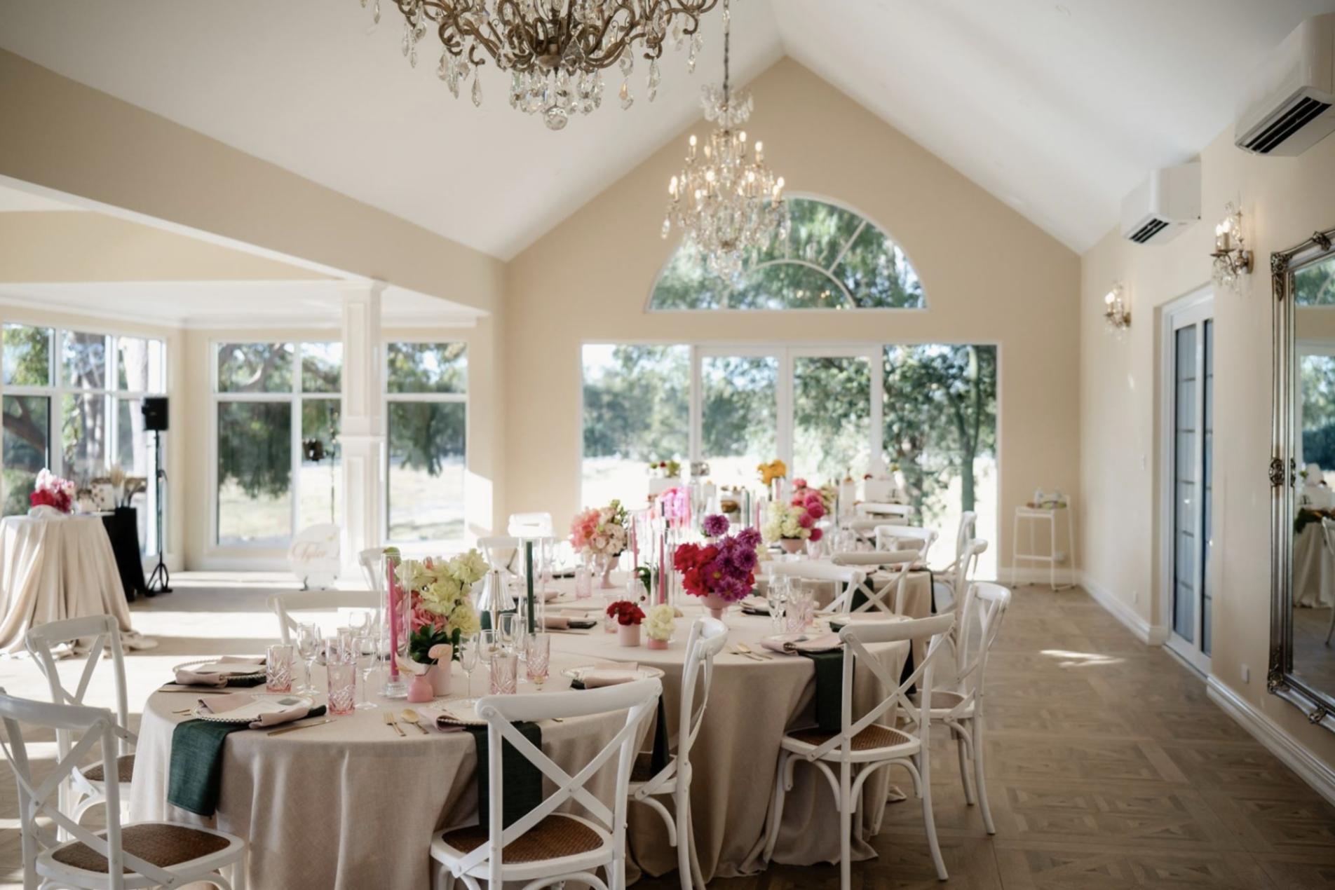 Interior of a bright, elegant event venue with high ceilings, large arched windows, and multiple chandeliers. The room is decorated with tables set for a celebration, featuring floral centerpieces, pink glassware, and white chairs. There are also air