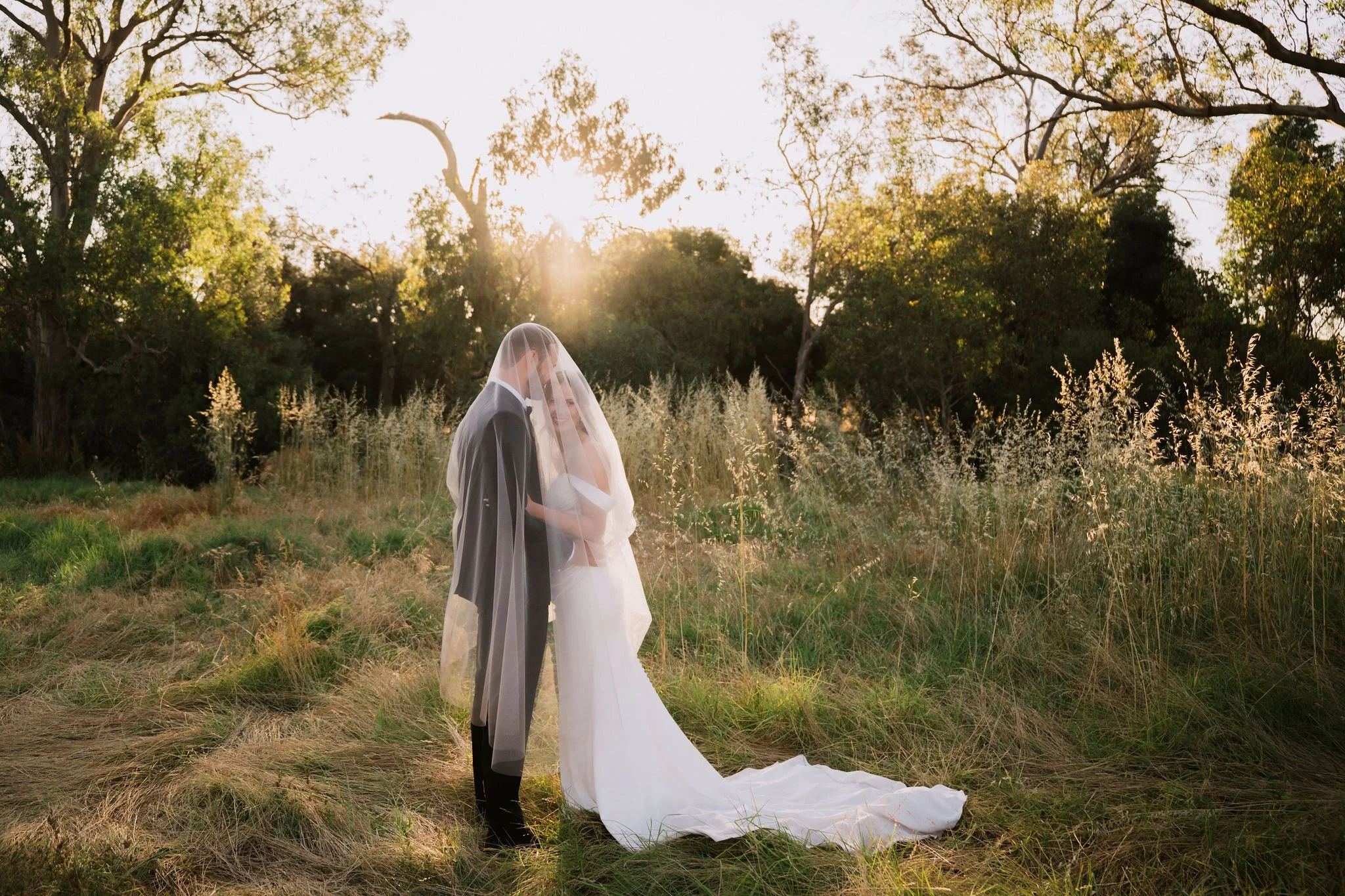 A bride and groom standing close together outdoors with a sunset in the background, groom in a black tuxedo and bride in a white wedding dress, covered by a veil.