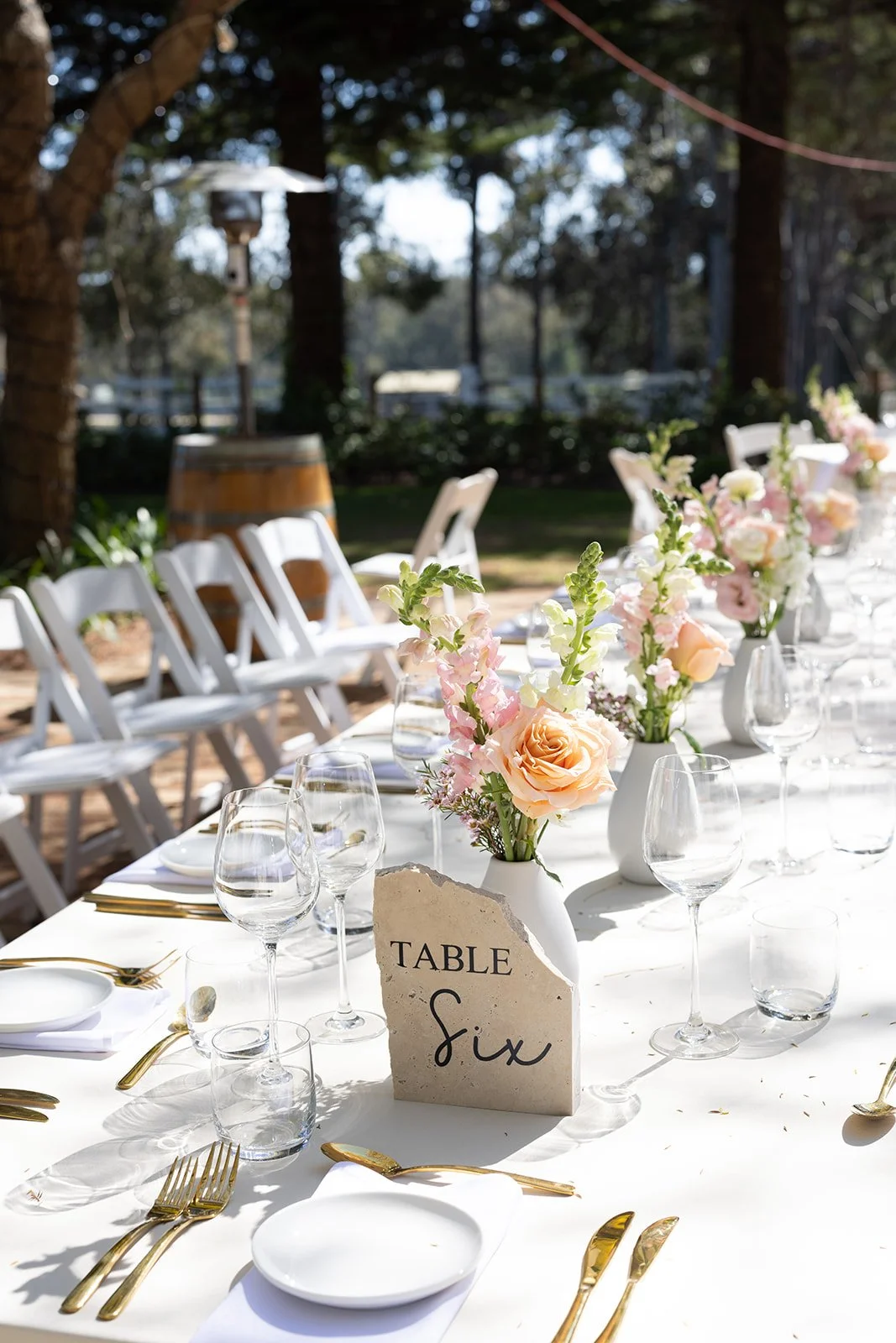 Wedding reception table setup outdoors with white tablecloth, elegant gold cutlery, wine glasses, white plates, and floral centerpieces in white vases, with a sign indicating "Table Six."