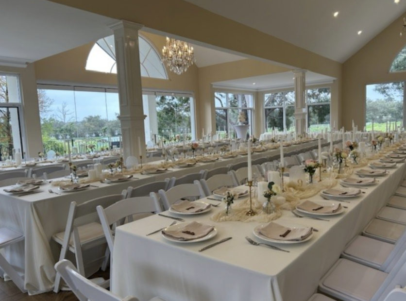 Elegant banquet setup in a bright, spacious dining room with large windows during daytime, featuring long tables with white tablecloths, floral centerpieces, and gold candlesticks.