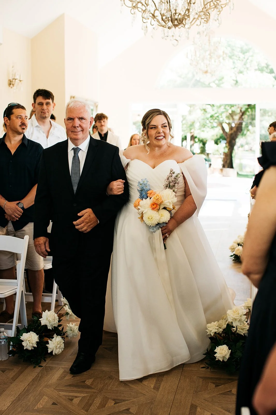 A bride in a white wedding gown holding a bouquet of flowers walking arm-in-arm with an older man, likely her father, during a wedding ceremony inside a brightly lit room with large windows and a chandelier.