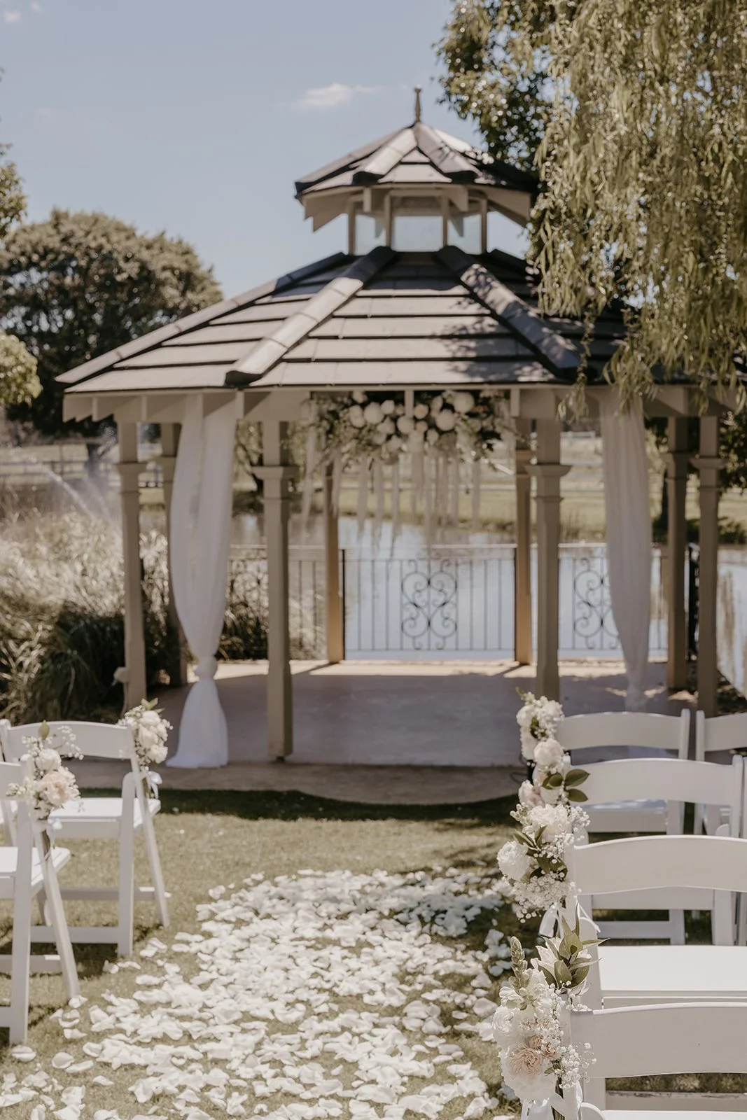 An outdoor wedding setup with a gazebo decorated with white curtains and floral arrangements. White chairs are arranged along a petal-lined pathway leading to the gazebo, with a scenic water background and trees.