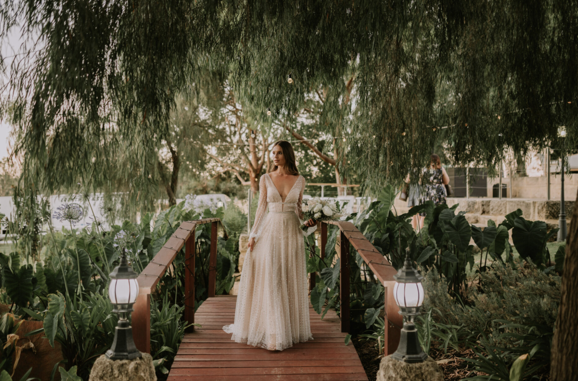 Bride in a cream-colored wedding gown holding a bouquet, standing on a small wooden bridge in a lush garden, with string lights hanging overhead and dense greenery surrounding her.