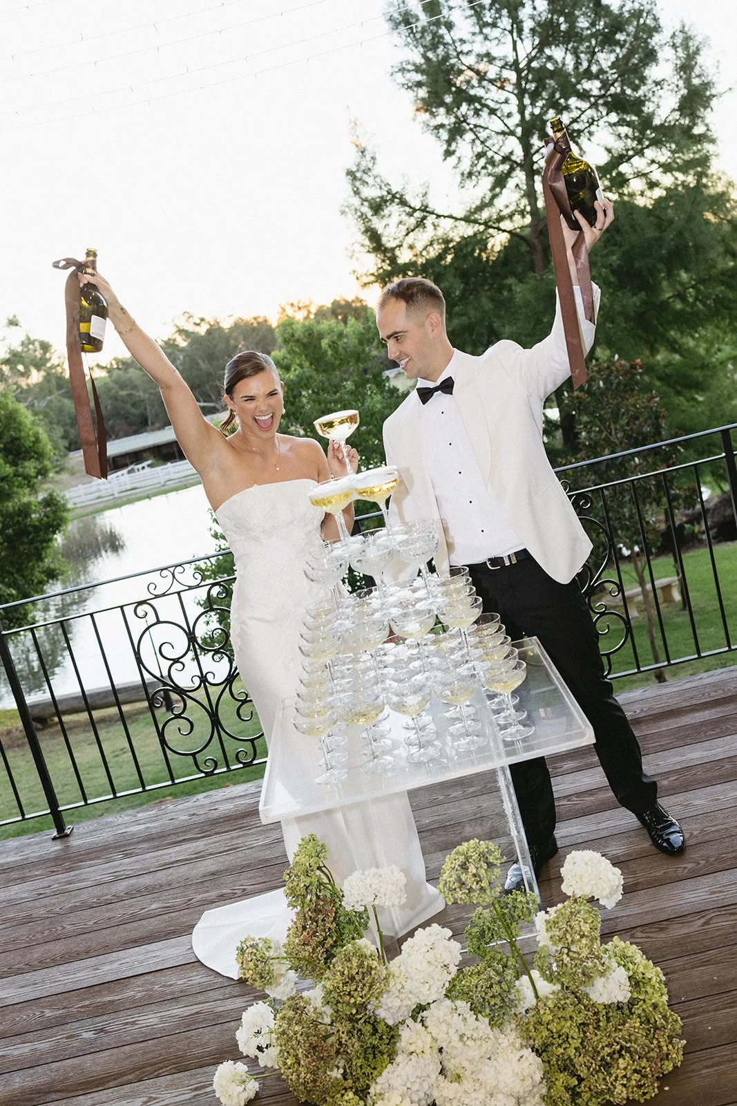 A bride and groom celebrating outdoors on a wooden deck with a table of champagne glasses. The bride, wearing a white strapless wedding dress, is holding a champagne bottle and smiling. The groom, in a white tuxedo jacket with black pants and bow tie