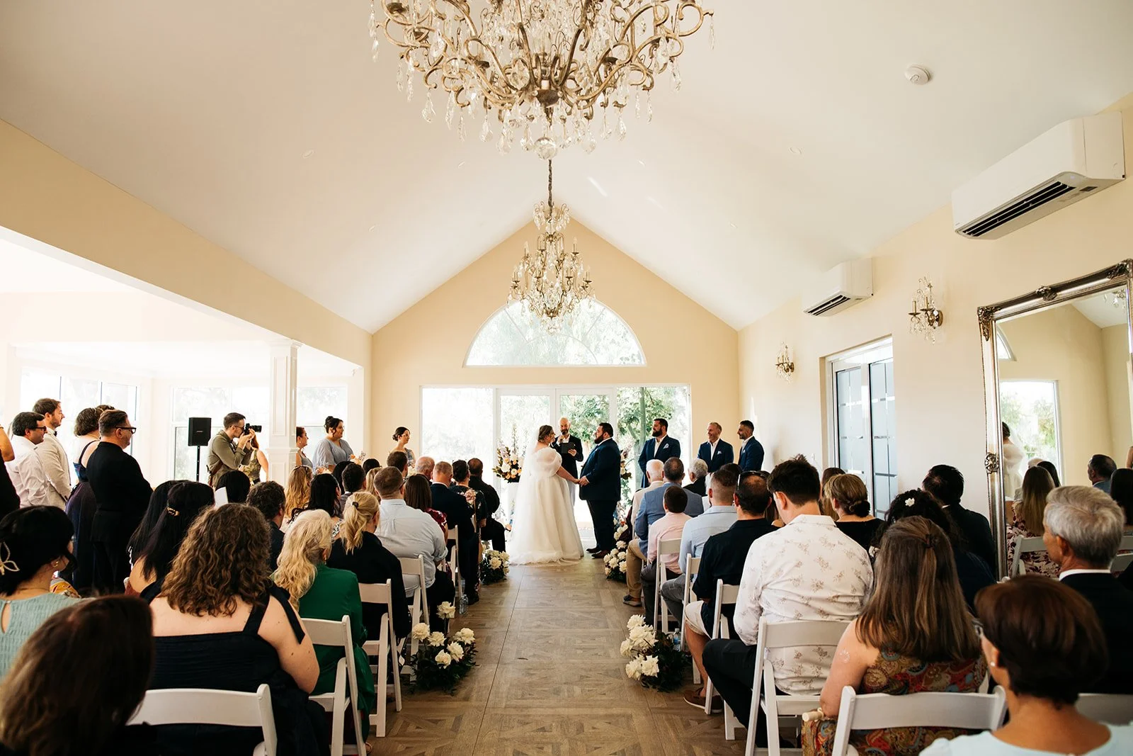 A wedding ceremony taking place inside a bright, elegant room with high ceilings, chandeliers, and large windows. The bride and groom are standing at the altar, holding hands, with officiant officiating. Guests are seated and standing around, watchin