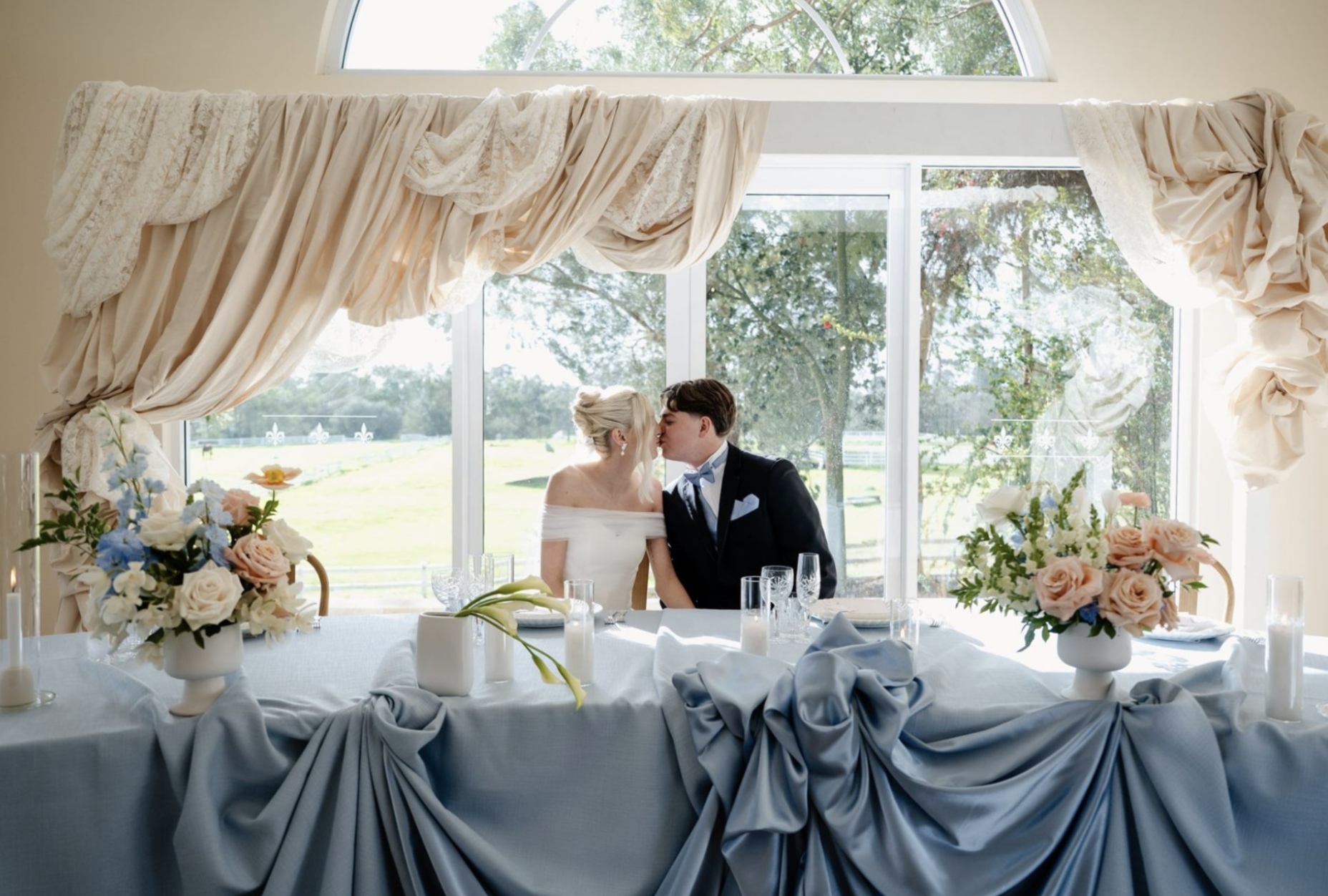 A bride and groom share a kiss at their wedding reception table, which is decorated with floral arrangements and draped tablecloth, in front of large windows showing a garden scene.