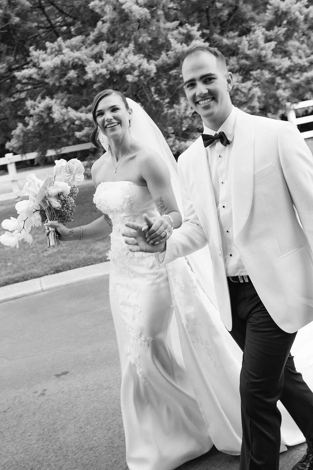 Black and white photo of a happy bride and groom walking outdoors, holding hands, with trees and a white fence in the background. The bride wears a strapless wedding gown and veil, holding a bouquet, and smiles. The groom is dressed in a tuxedo with 