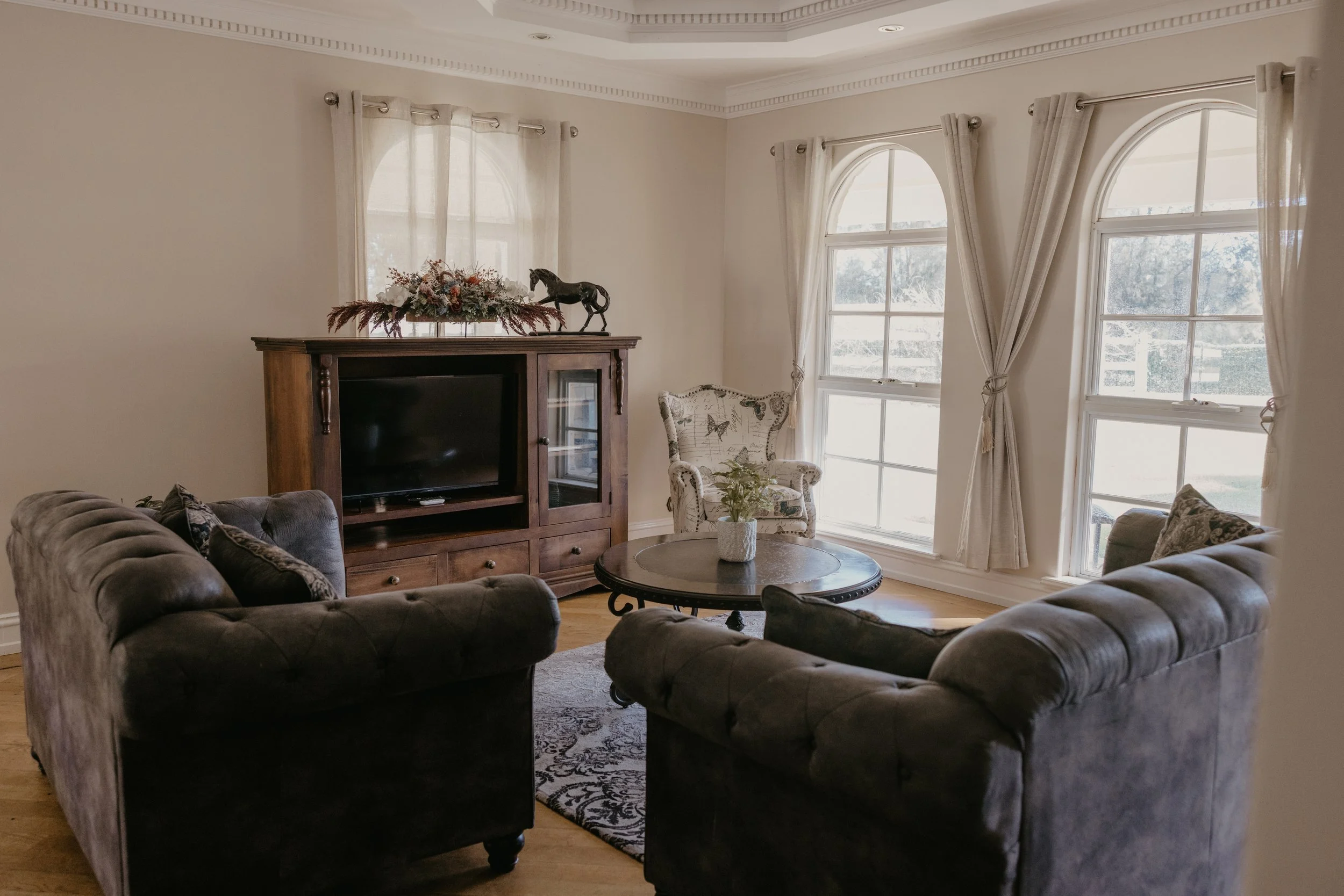A living room with large arched windows covered with light-colored curtains, a dark sofa set, a round coffee table with a vase of flowers, a patterned armchair, and a wooden cabinet with a decorative horse and floral arrangement on top.