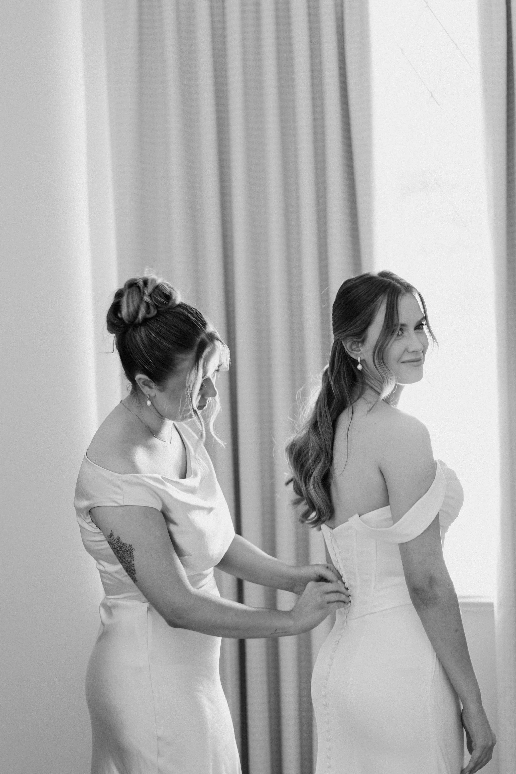 A bride in an off-shoulder wedding dress looks over her shoulder as a woman helps her button up the back of her gown, standing in front of curtains.