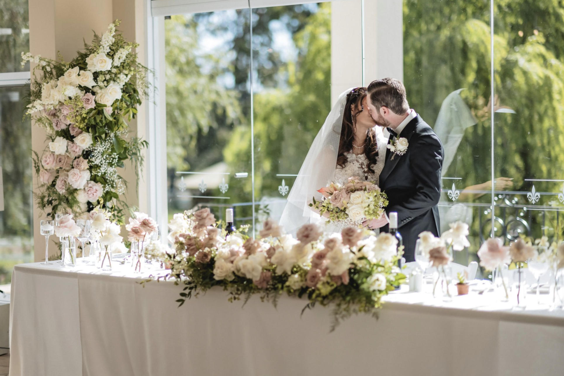 A bride and groom kissing during their wedding ceremony inside a bright room with large windows, decorated with white and light pink flowers and greenery, with a table full of floral arrangements and Champagne glasses in front of them.