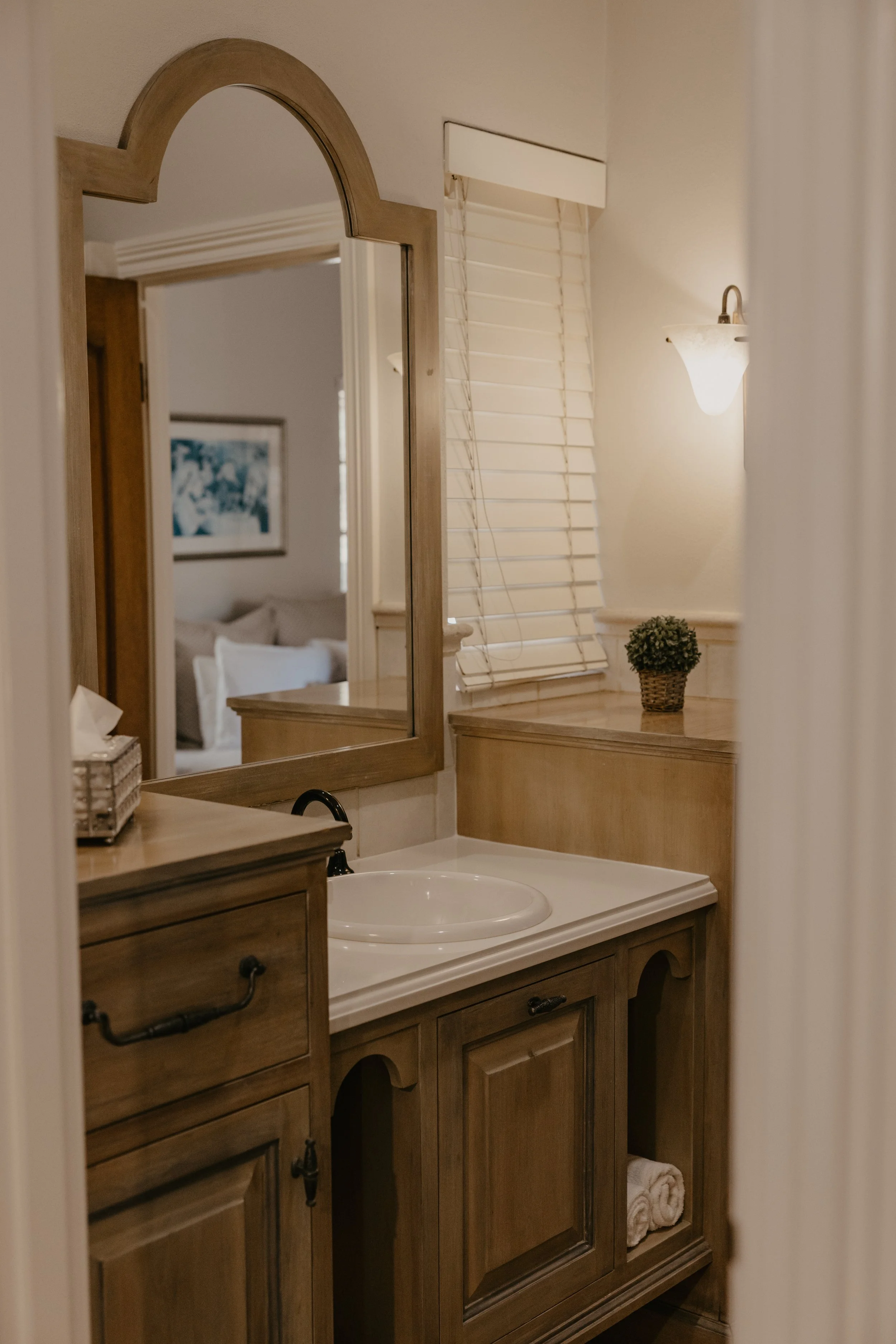 A bathroom vanity with a wooden cabinet and a white sink, a mirror with a wooden frame, a window with white blinds, a small potted plant, a wall-mounted light fixture, and a glimpse of a bedroom in the background.