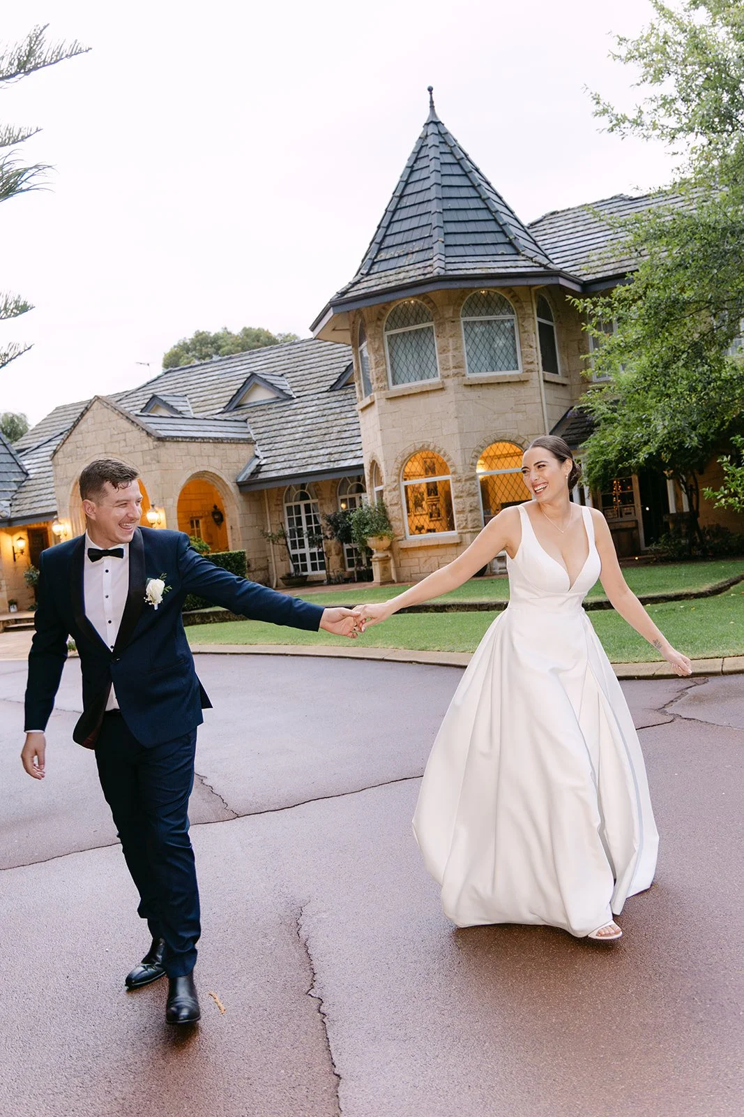 A bride and groom holding hands and smiling outside a large house with stone walls, arched windows, and a tower, during their wedding celebration.