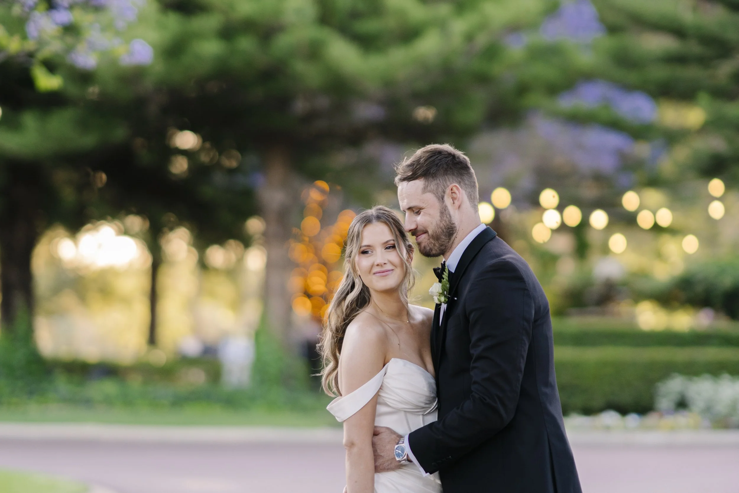 A bride and groom sharing a moment outdoors during sunset with blurred trees and string lights in the background.