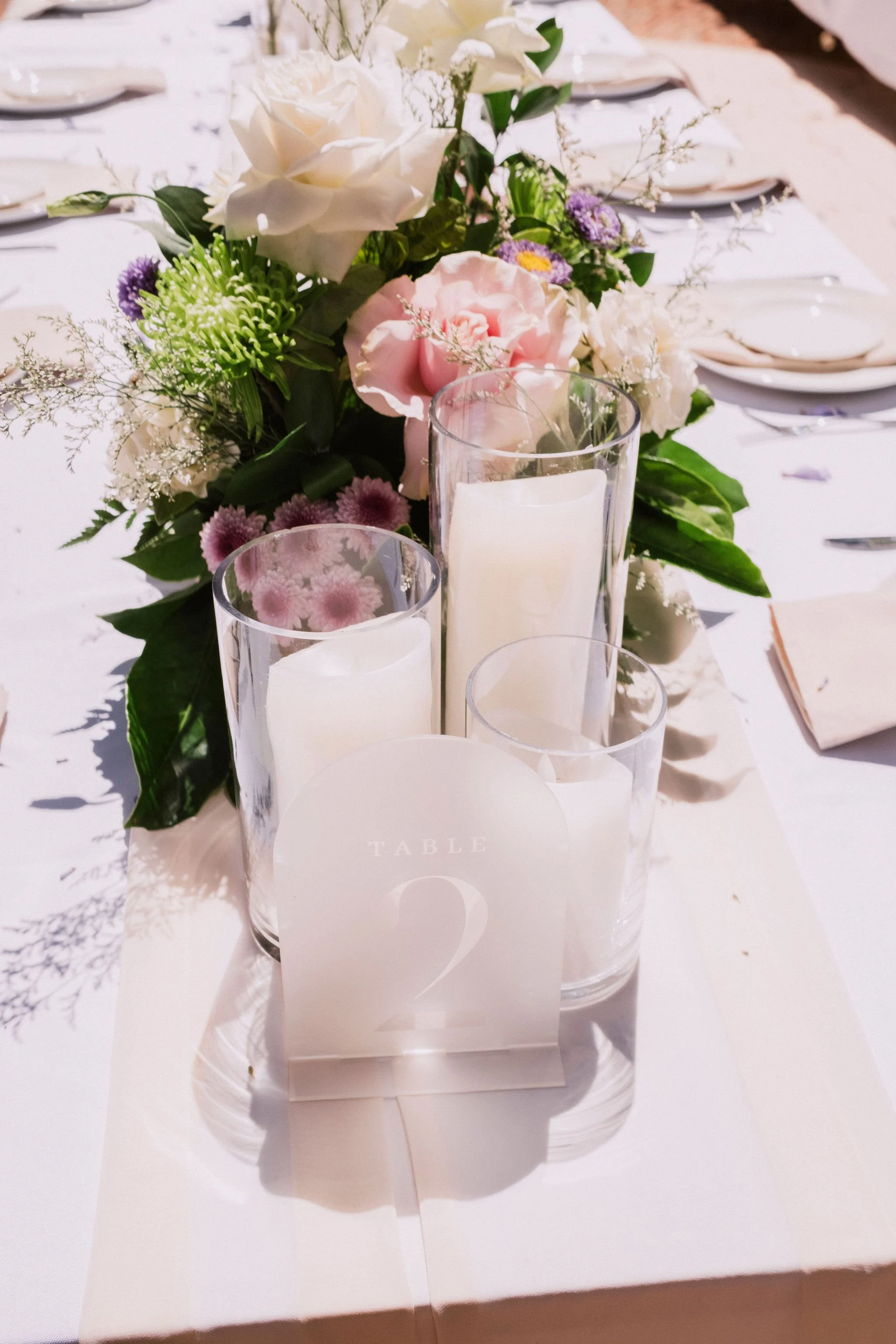 A table centerpiece with white and pink roses, purple, and green flowers, along with three white candles in glass holders, on a white tablecloth at a formal event or wedding reception.