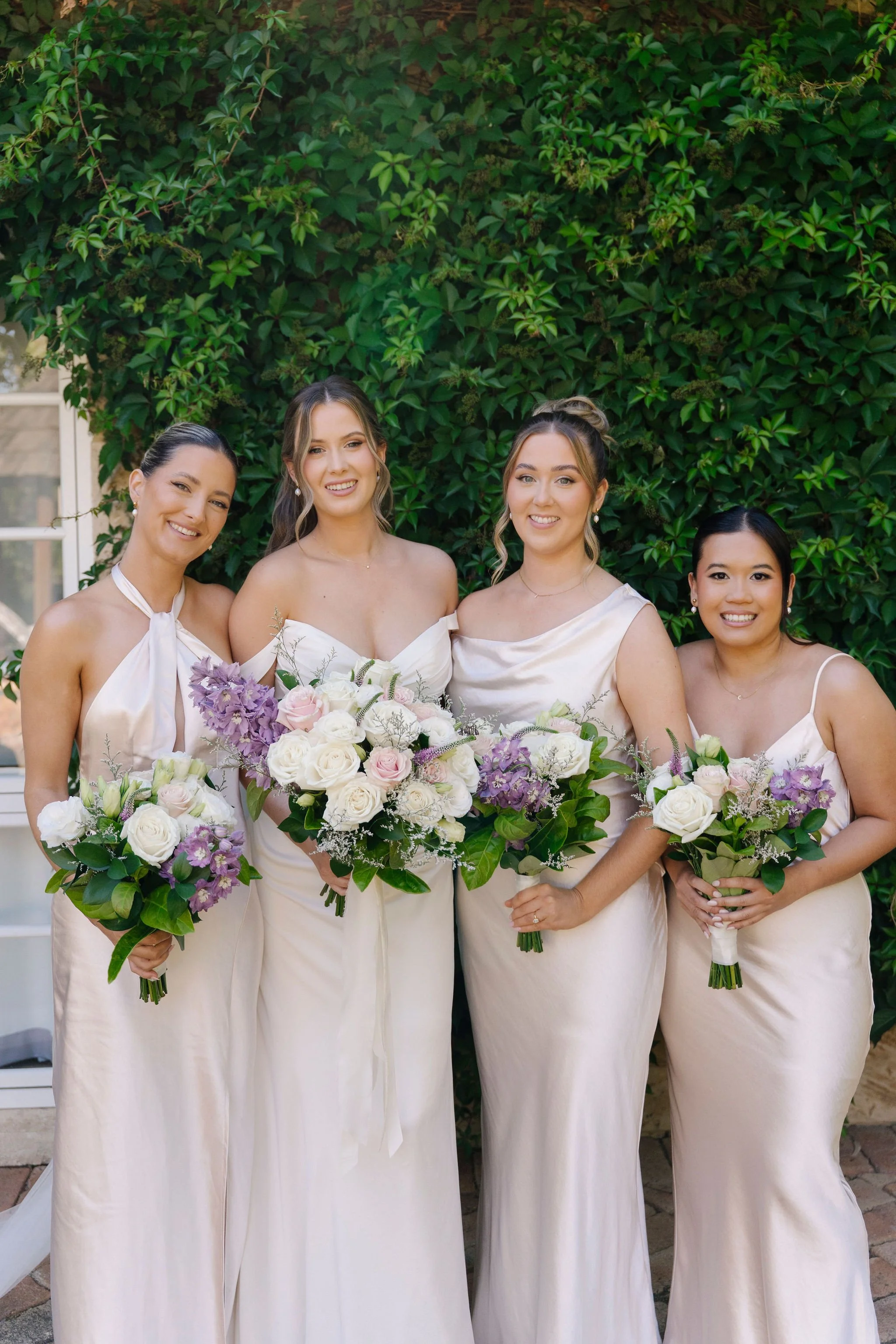 Four women in elegant dresses holding bouquets of flowers, standing outdoors in front of a lush green hedge, smiling at the camera.