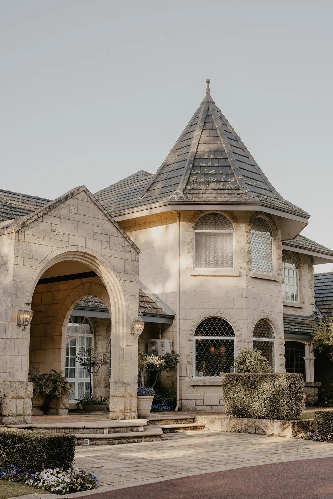 A large house with stone exterior walls, arched windows with diamond-shaped panes, a rounded tower with a conical roof, and a front porch with potted plants and shrubs.