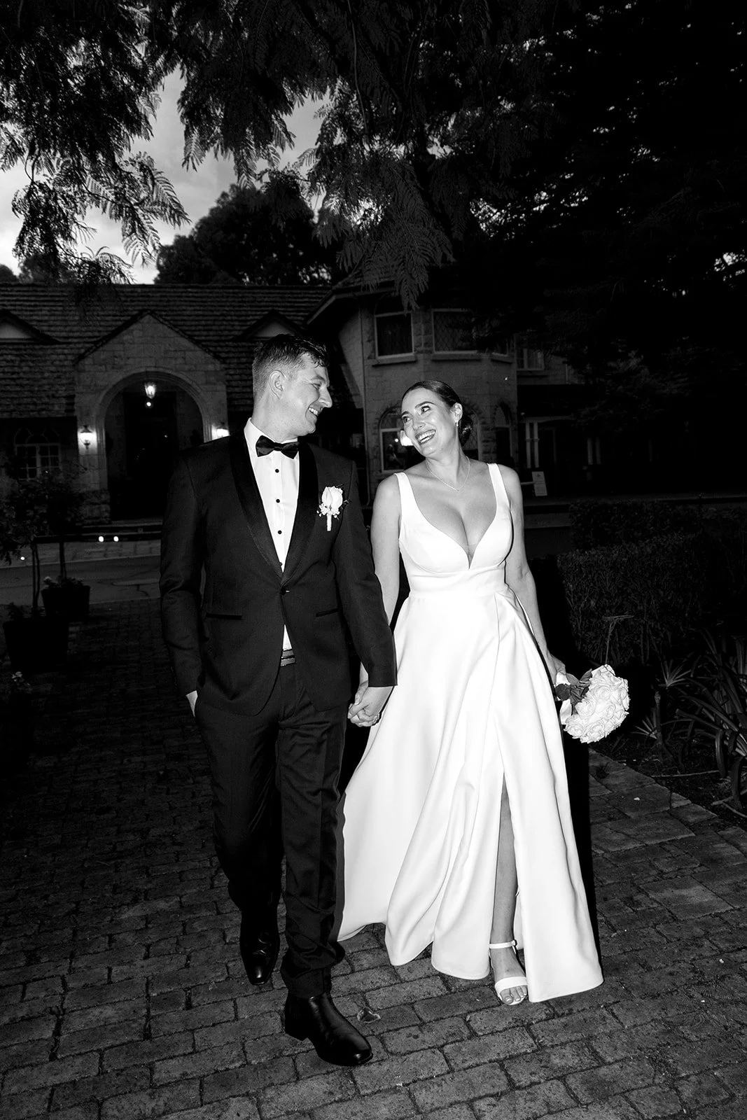 A black and white photo of a newly married couple walking arm in arm outside at night. The groom is wearing a black tuxedo with a bow tie, and the bride is in a white wedding gown holding a bouquet of flowers. They are smiling and looking at each oth