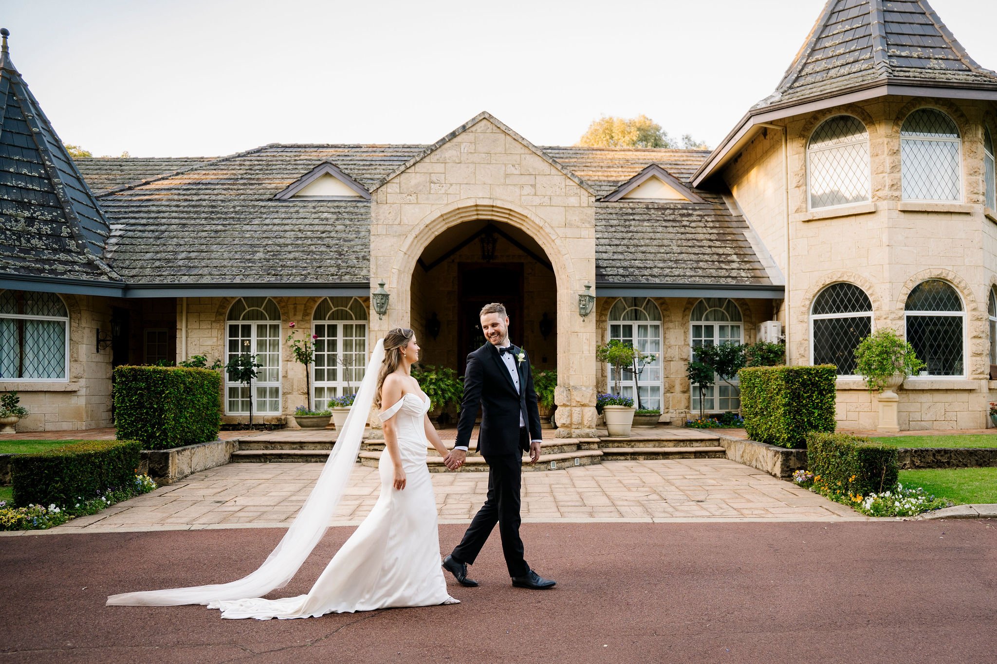 Bride and groom holding hands, walking in front of a large stone house, each smiling at the other.