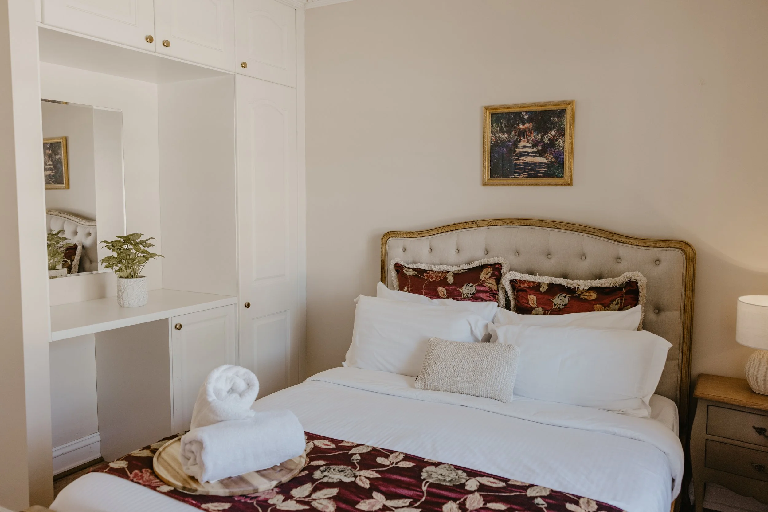 A neatly made bed with white linens and decorative pillows, a bedside table with a lamp, and a framed picture of a garden with a pathway on the wall behind it, in a cozy bedroom.