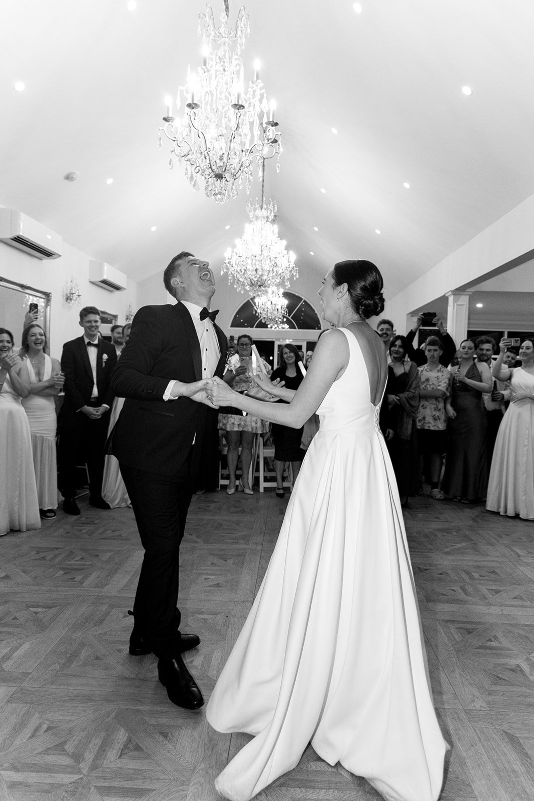 A bride and groom sharing a dance at their wedding reception, surrounded by smiling guests in a decorated ballroom with chandeliers.