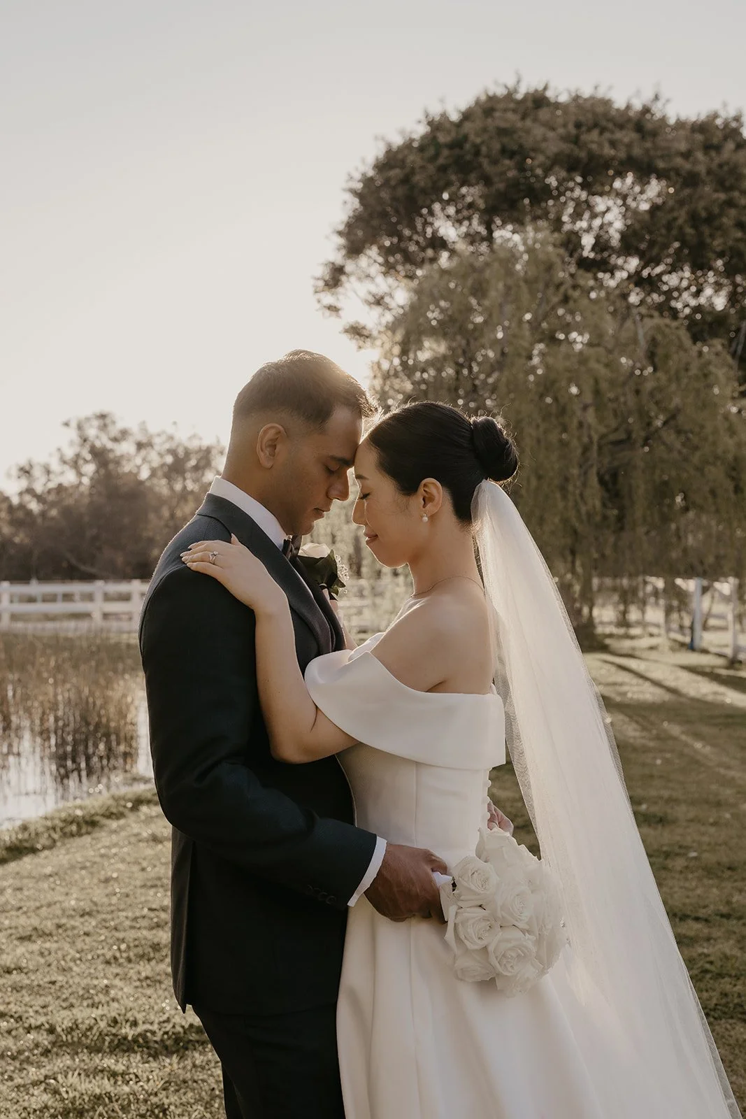 A bride and groom standing close with foreheads touching, outdoors near a pond and trees, during sunset, in wedding attire, holding a bouquet of white roses.