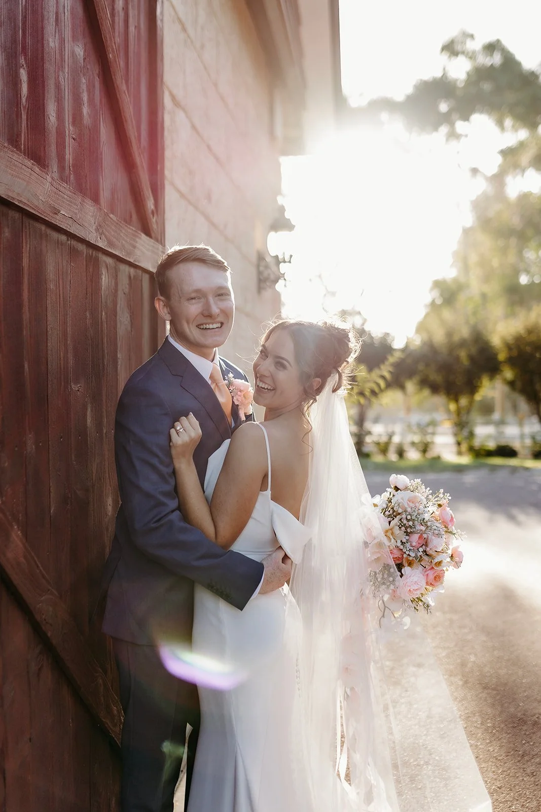 A newlywed couple smiling and laughing outdoors at sunset, standing against a wooden wall. The bride holds a bouquet of pink and white flowers, and the groom is dressed in a navy suit with a pink boutonniere. Bright sunlight creates a warm glow.