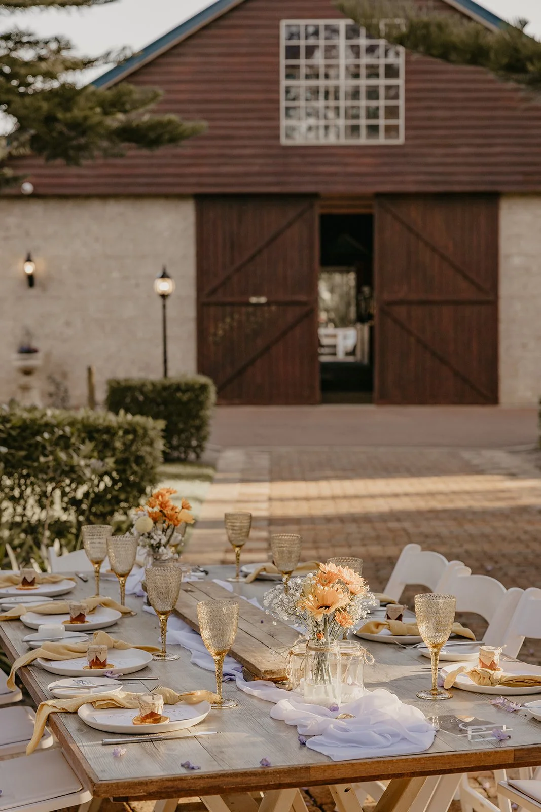 Outdoor long table set for a wedding celebration at Brookleigh Estate Perth with floral centerpieces, gold glasses, plates, and napkins, in front of a barn-style building during daytime.