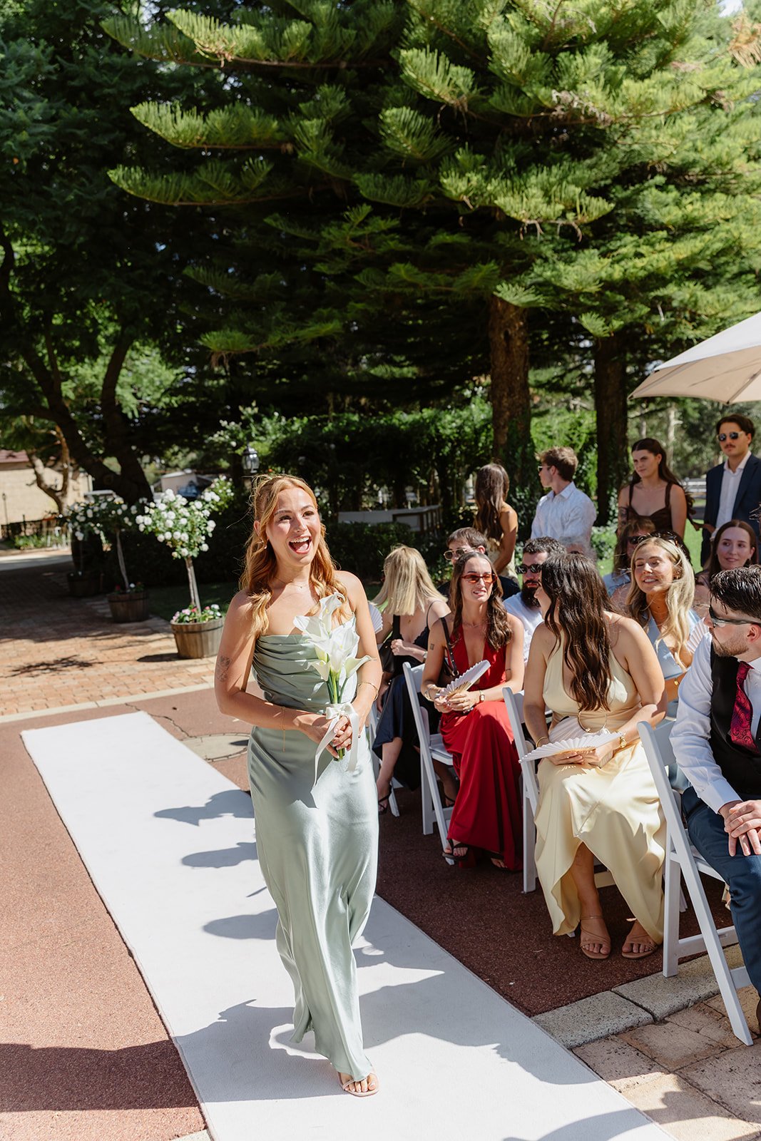 A woman in a light green satin dress walking down an outdoor aisle at a wedding, holding a white calla lily bouquet, with guests seated on white chairs on either side under trees.