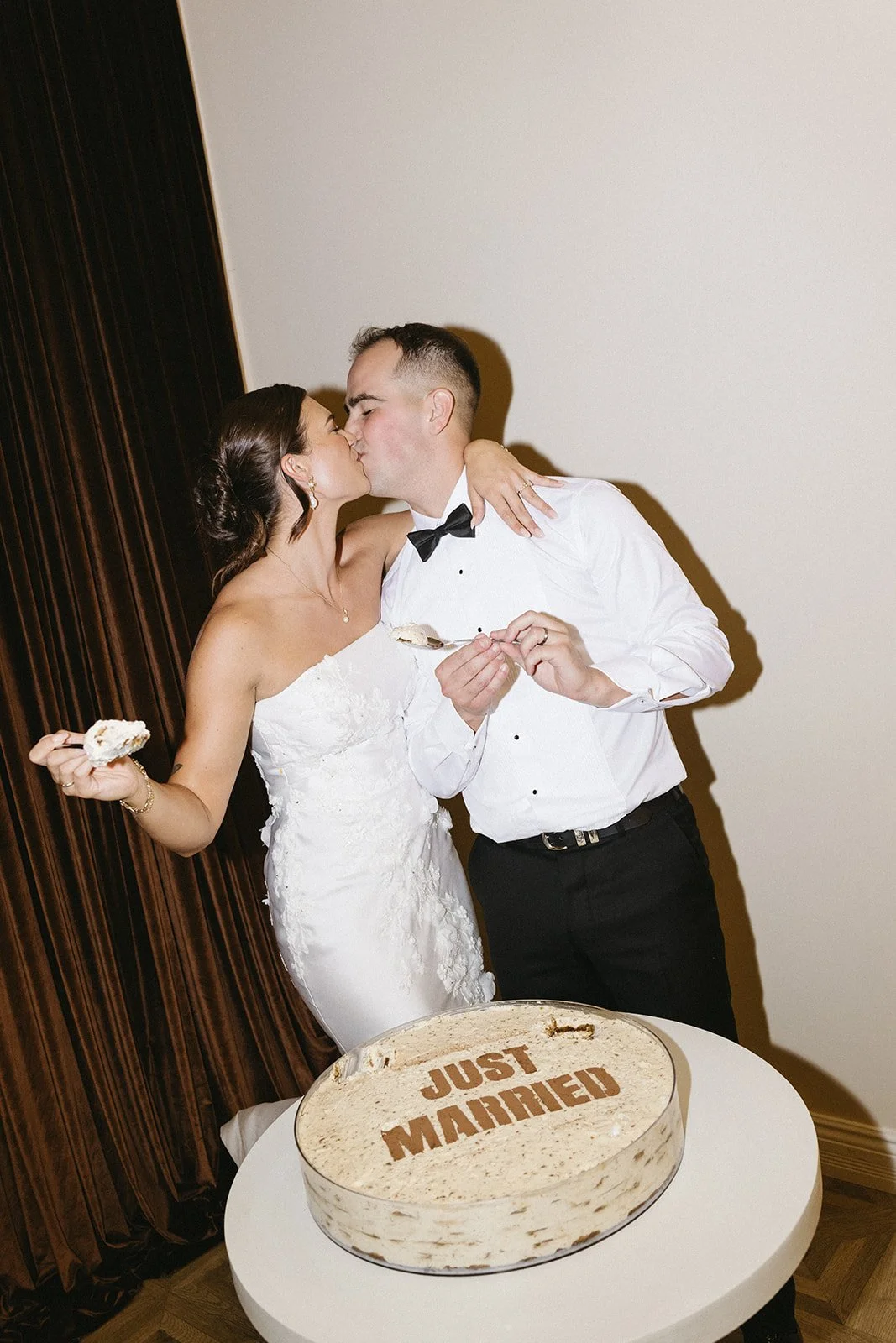 A newlywed couple kissing at their wedding reception, with a cake in front that says 'Just Married'.