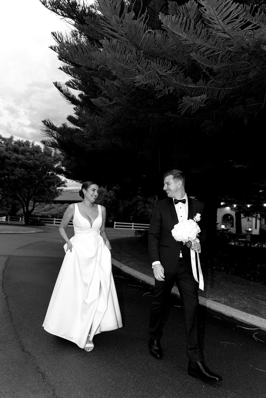 Black and white photo of a bride and groom walking outdoors, smiling at each other. The bride is wearing a wedding dress and the groom is in a tuxedo holding a bouquet of flowers.