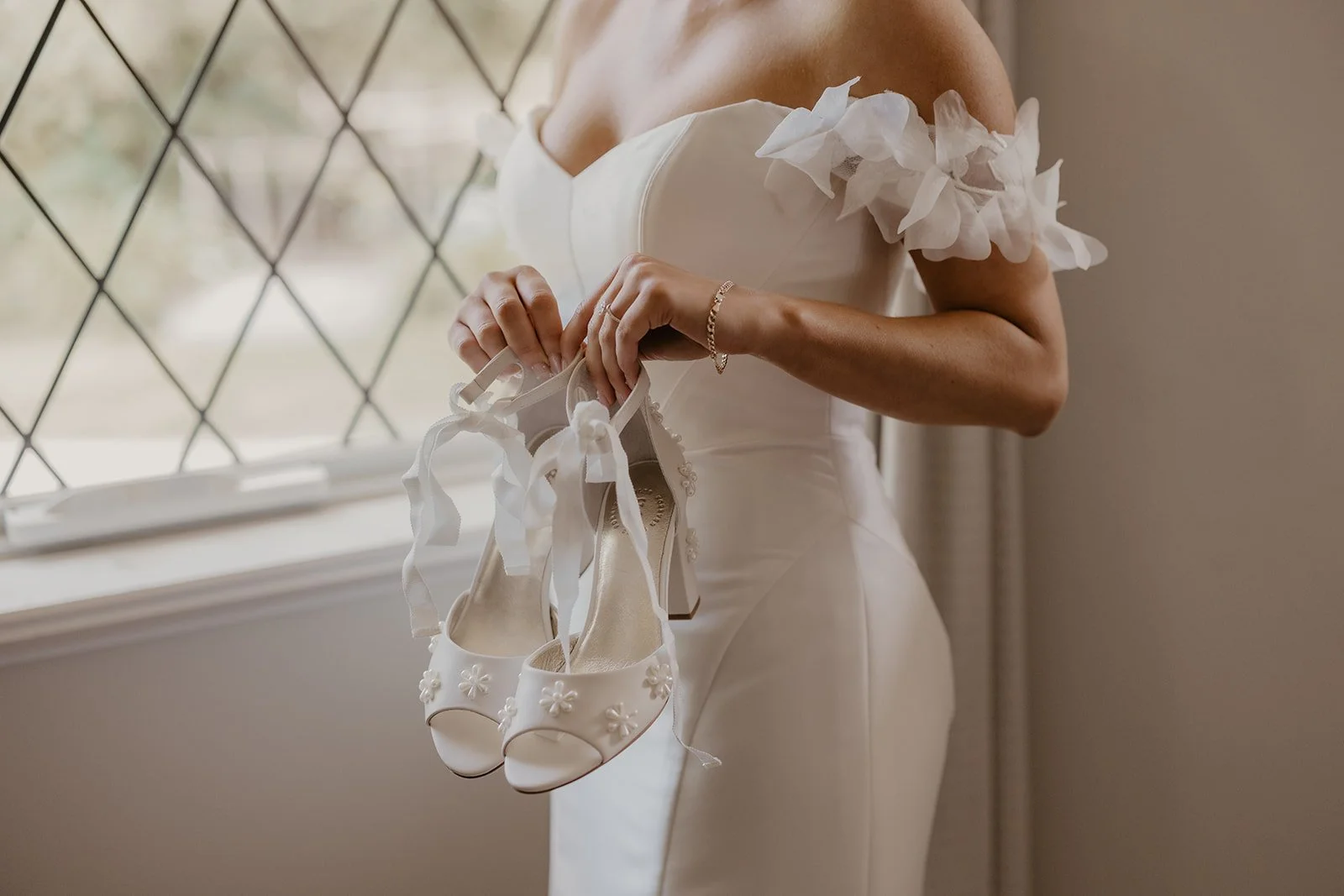 A woman in a white wedding dress holding white wedding shoes with floral embellishments and ribbon ties.