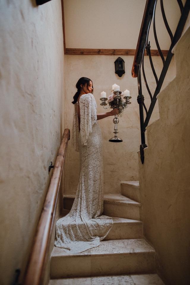 A woman in a long, elegant white lace dress stands on a staircase, holding a vintage candelabra with lit candles, in a rustic interior setting.