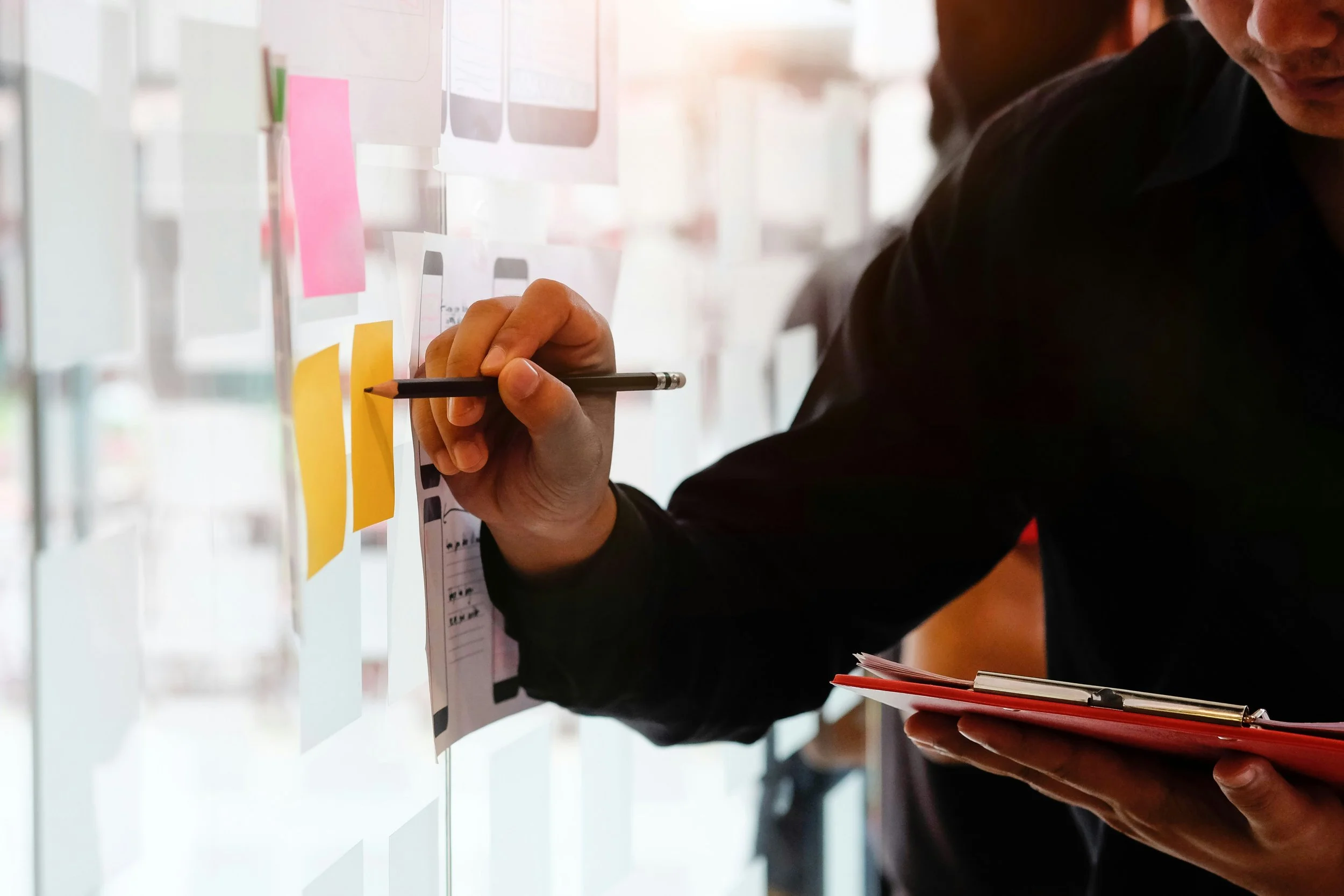 Person attaching sticky notes to a glass wall with other notes and papers in the background.