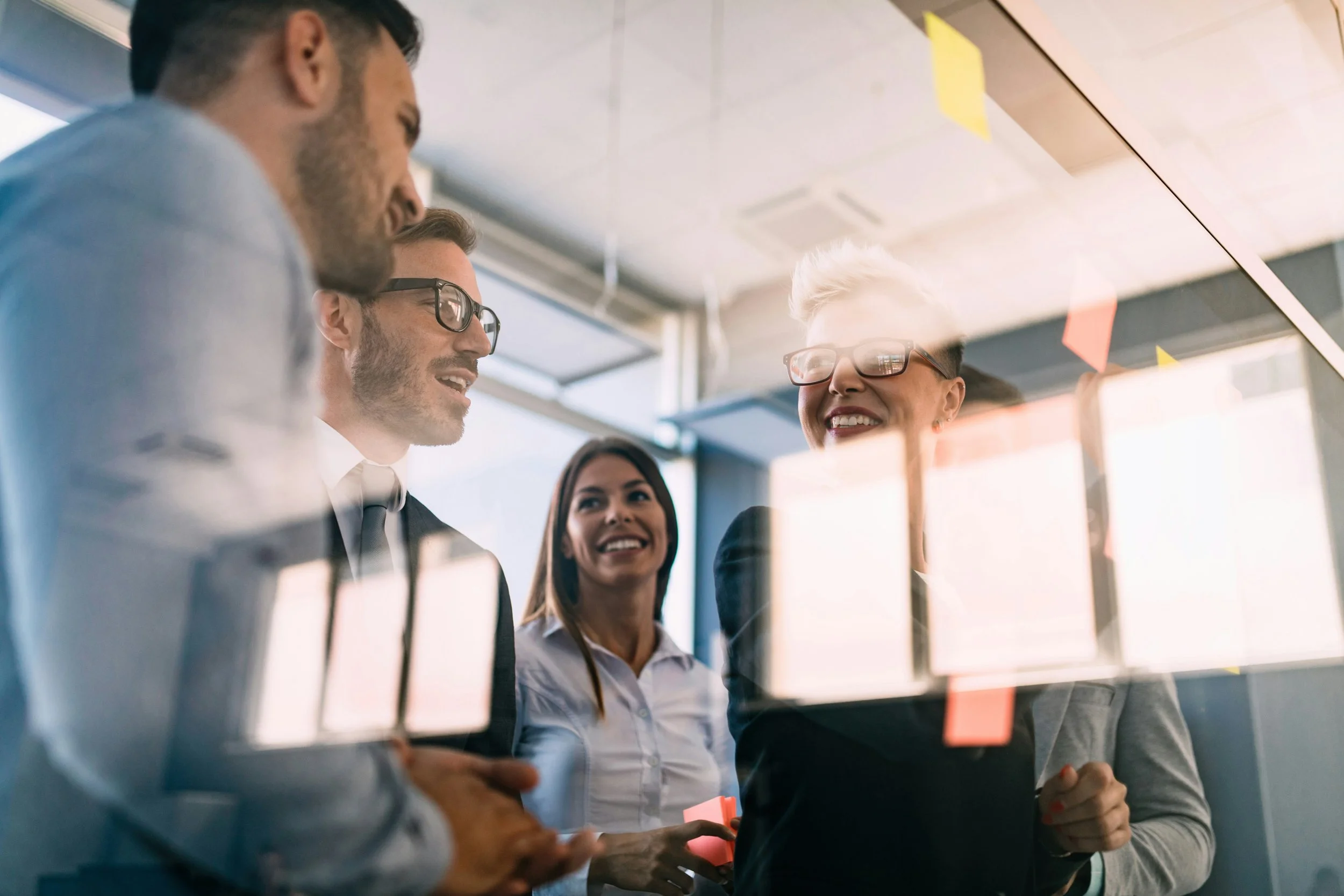 Four business professionals, two men and two women, gathered around a glass wall with sticky notes, discussing and smiling in a modern office.