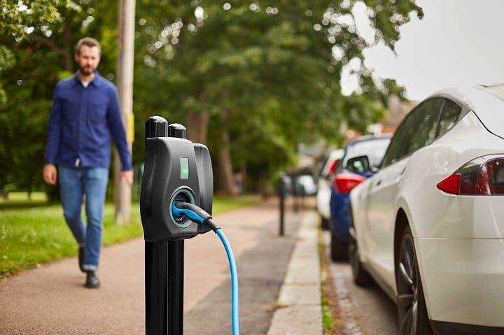A man walking on a sidewalk past an electric vehicle charging station connected to a white car parked on the street.