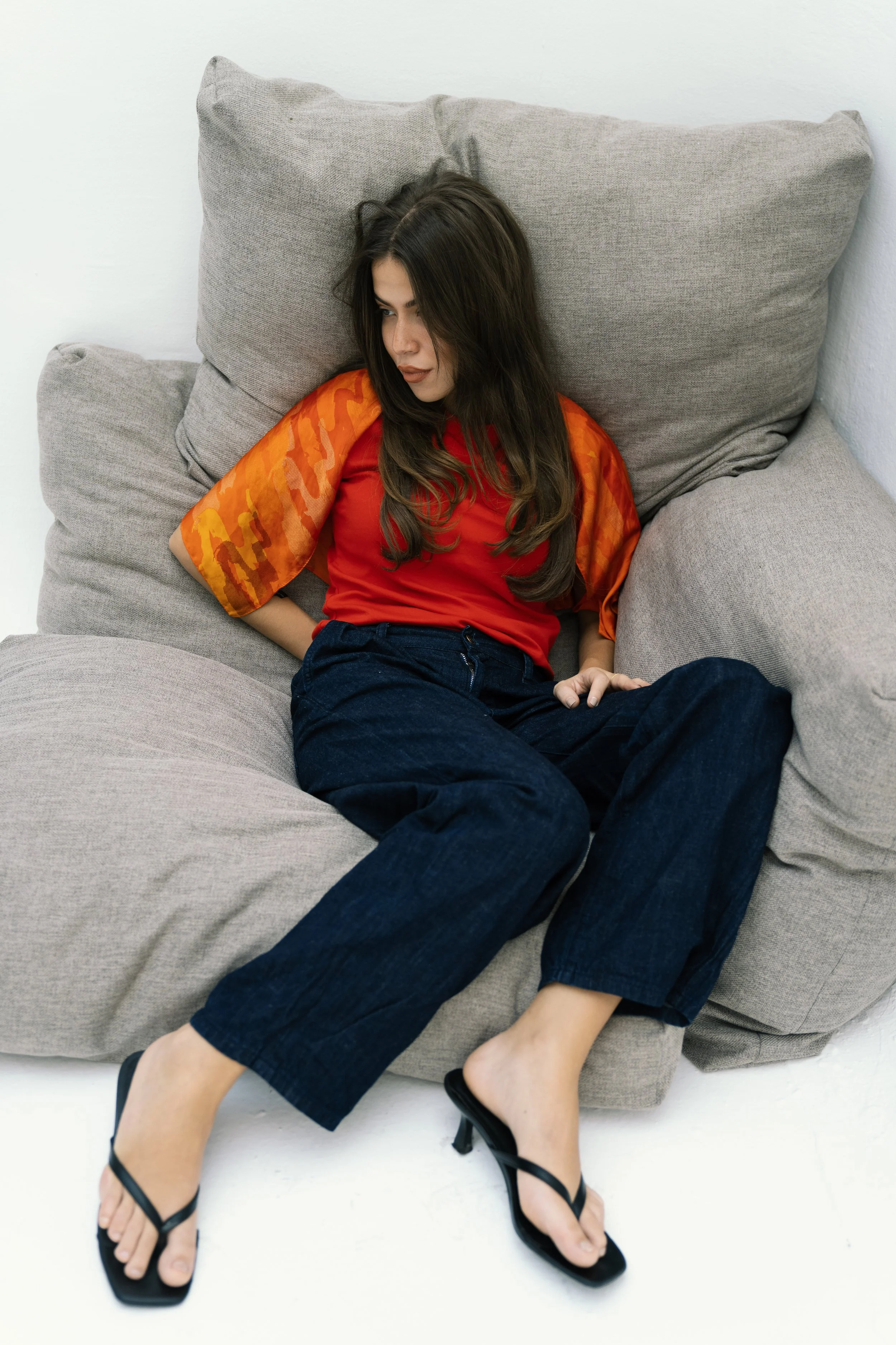 A woman with long dark hair wearing a red top with orange patterned sleeves, dark jeans, and black heeled sandals sitting on a light gray sofa, leaning against cushions, looking down with a relaxed pose.