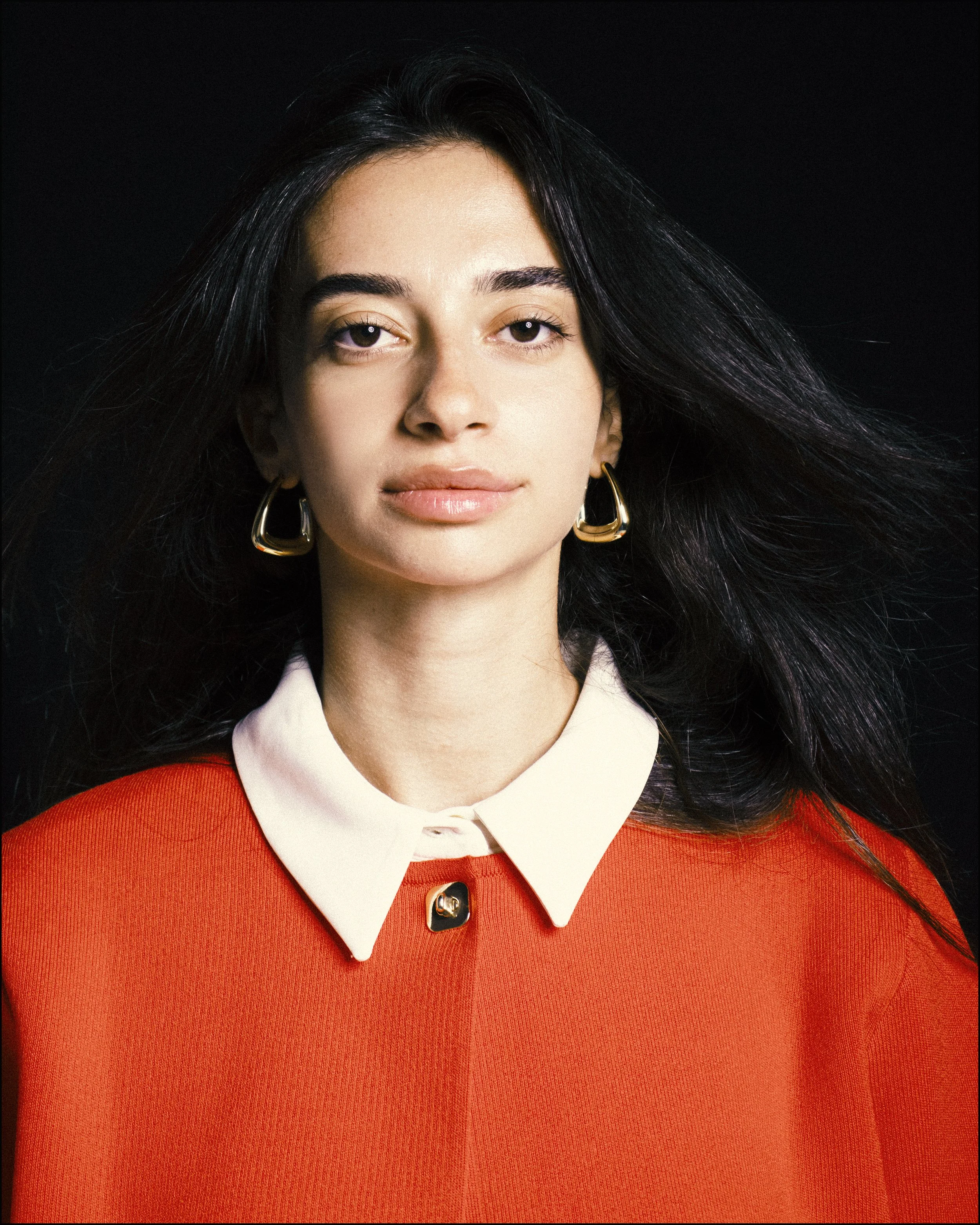 A portrait of a young woman with long dark hair wearing a red top with a white collar and gold earrings, against a black background.